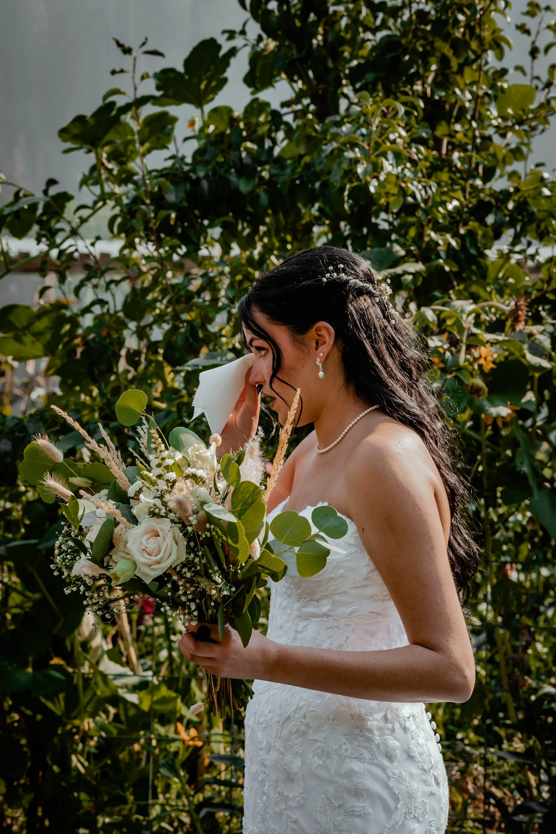 A bride in a white wedding dress with black hair holding a bouquet of white roses and greenery, with her eyes closed and touching her face, standing outdoors among green bushes.