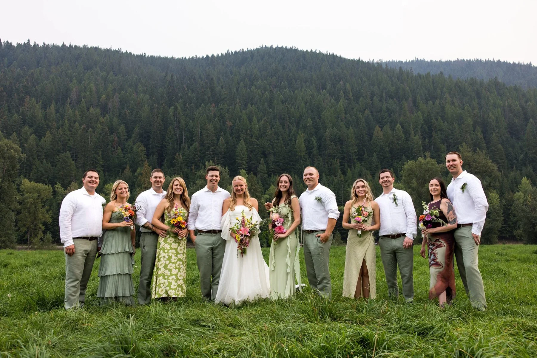 Group of people in a wedding photo standing in a grassy field with a forested hillside in the background, some holding bouquets of flowers.