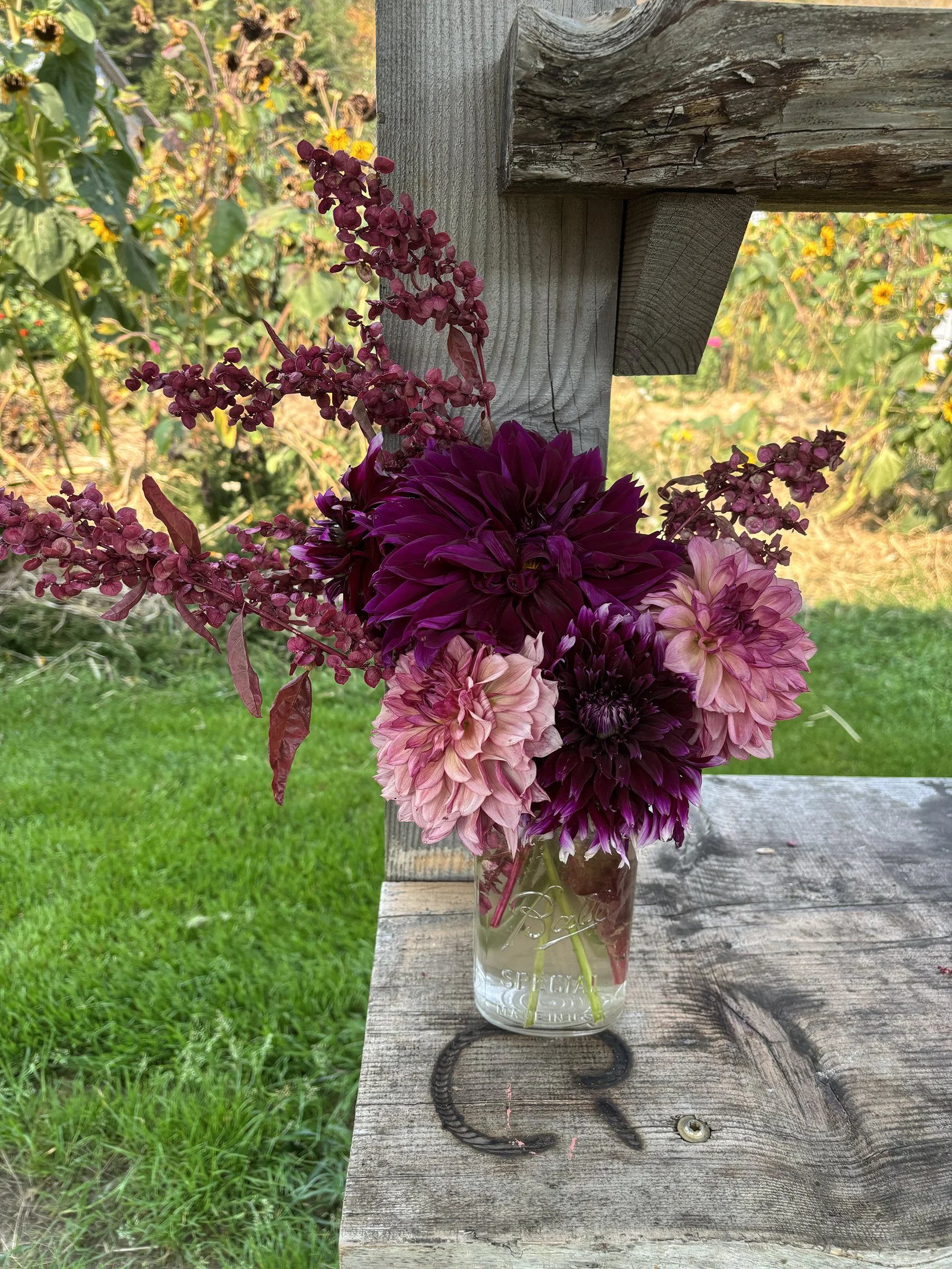 A bouquet of pink and dark purple flowers in a glass jar on a weathered wooden surface outdoors.