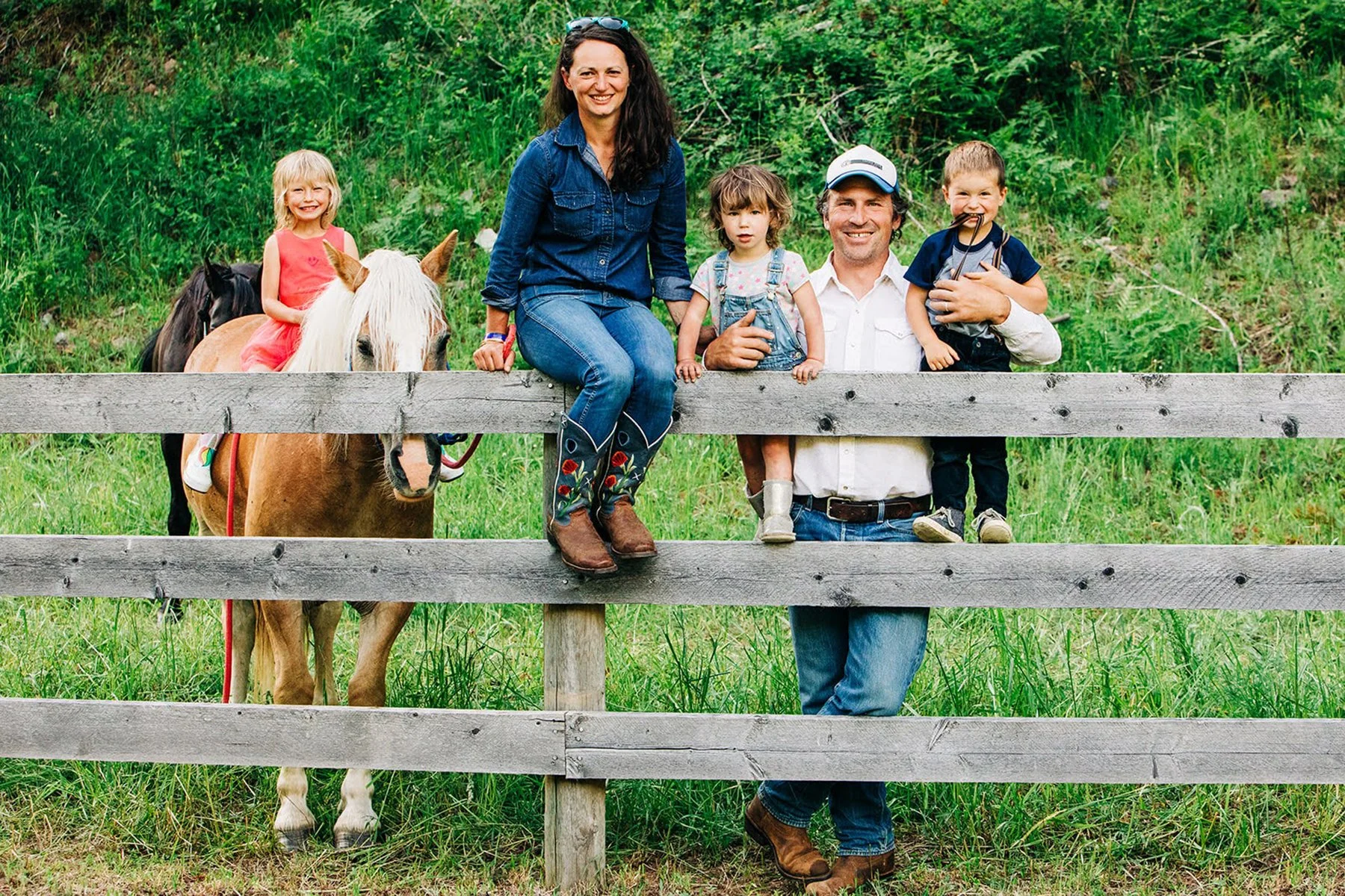 A family of five with two children and two adults, along with a horse, standing behind a wooden fence in a green outdoor setting.
