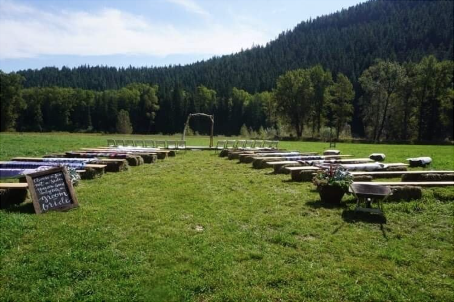 Outdoor wedding setup on a grassy field with wooden benches arranged in rows, a blackboard sign, and mountain and forest in the background.