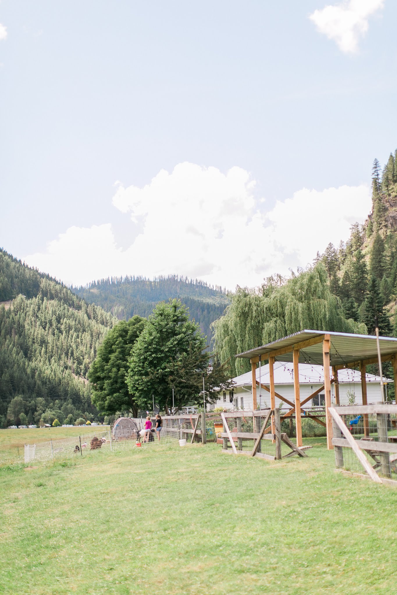A peaceful farm scene with a grassy field, trees, a building with a covered porch, and a backdrop of mountains and sky.