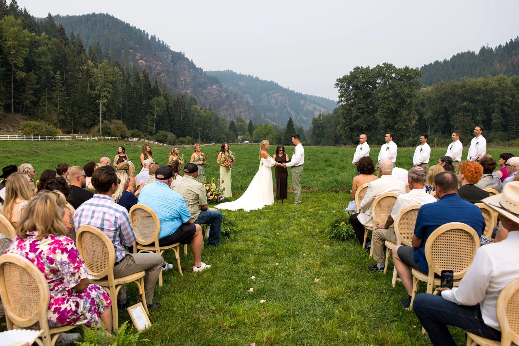 Outdoor wedding ceremony in a lush green field with mountains and trees in the background, bride and groom exchanging vows, surrounded by guests seated on chairs.