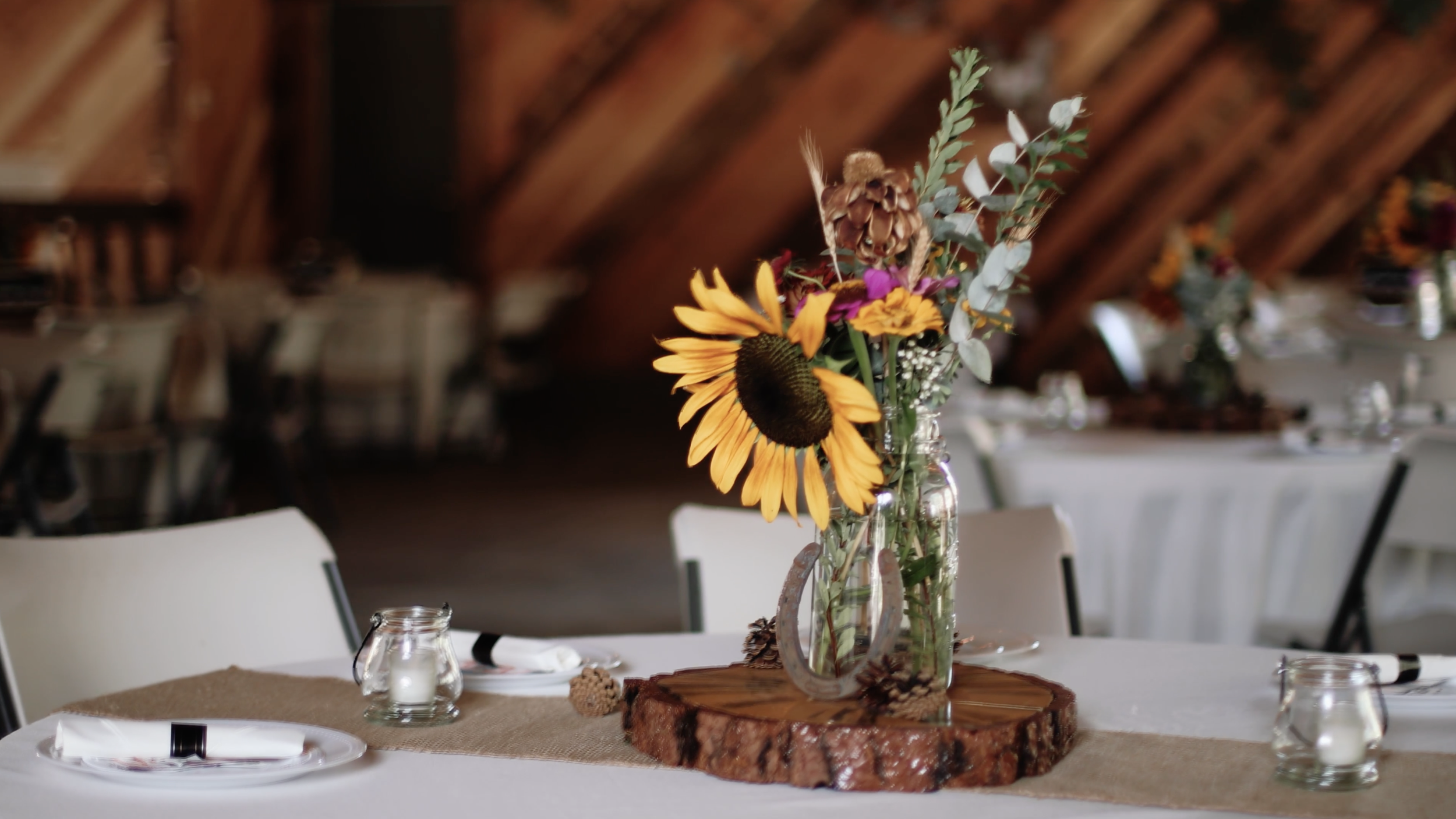 A rustic table centerpiece featuring a glass jar filled with a bouquet of dried and fresh flowers, including a sunflower, placed on a wooden slab in a decorated event space.