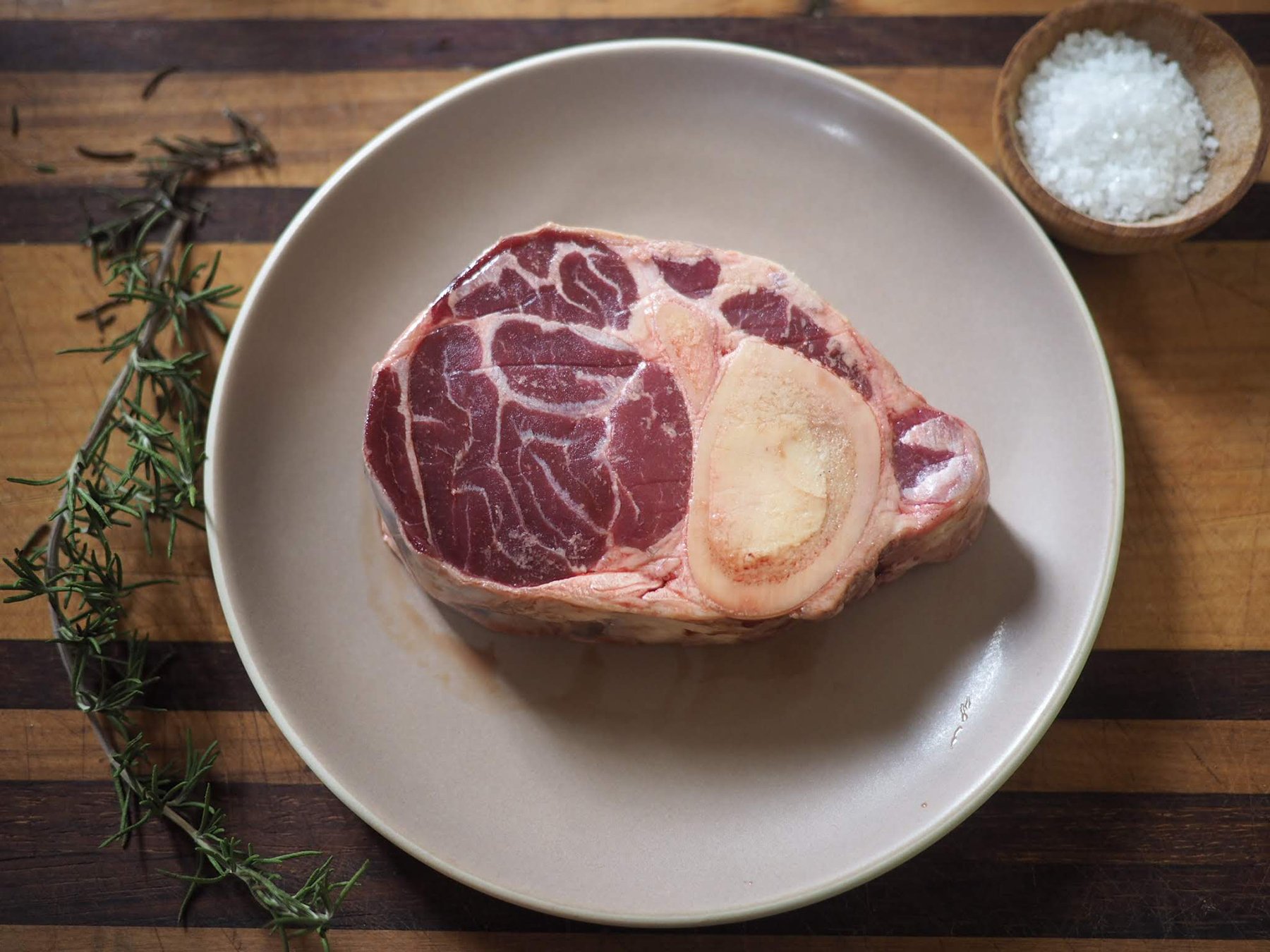 A raw beef shank with bone on a gray plate. There is a small bowl of coarse salt and a sprig of fresh rosemary on a wooden surface.