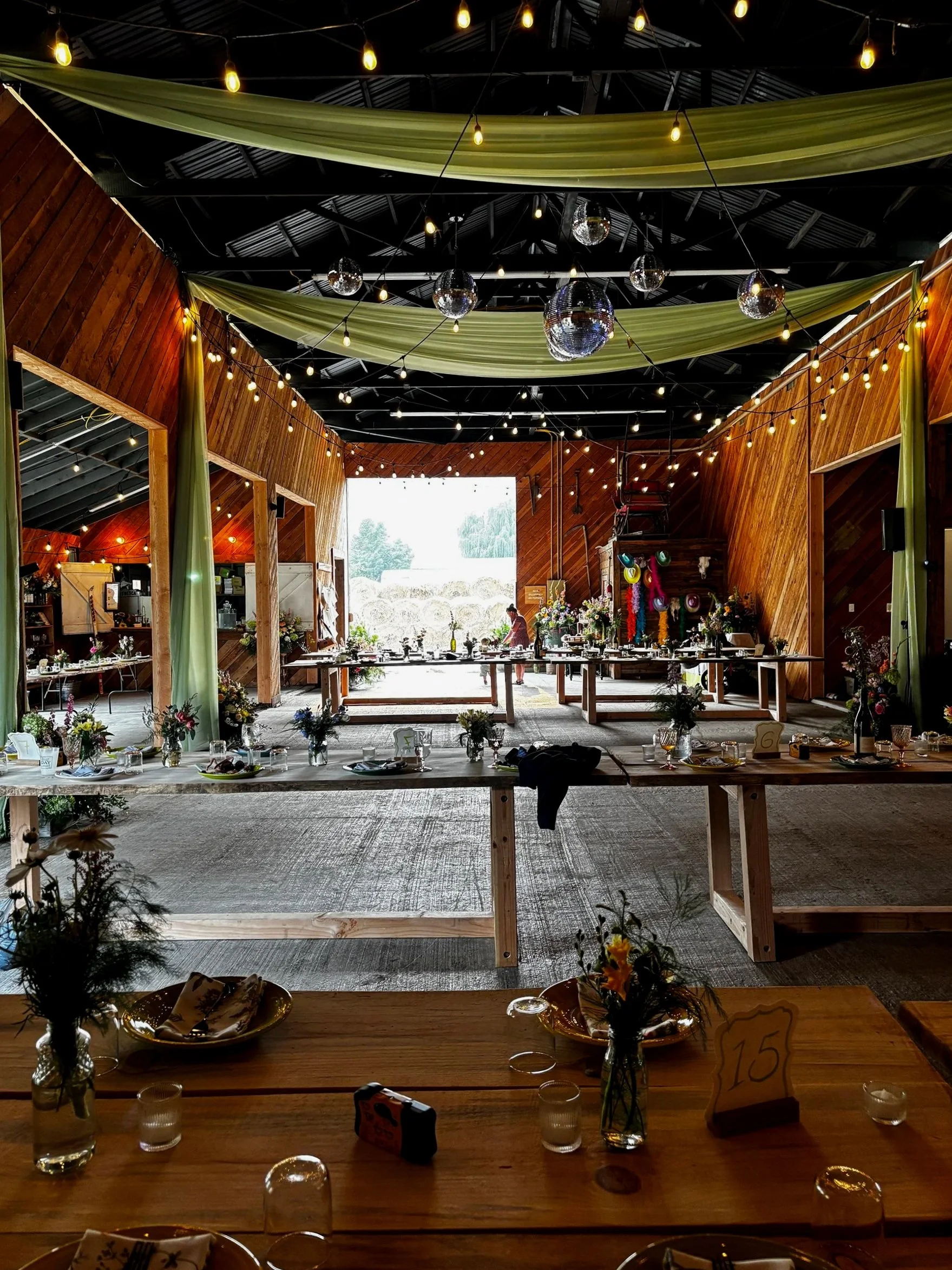 Indoor wedding reception with wooden walls and ceiling, decorated with string lights, disco balls, and green fabric drapes. Long tables with floral centerpieces, glassware, and place settings are arranged for guests, with an open barn-style entrance 