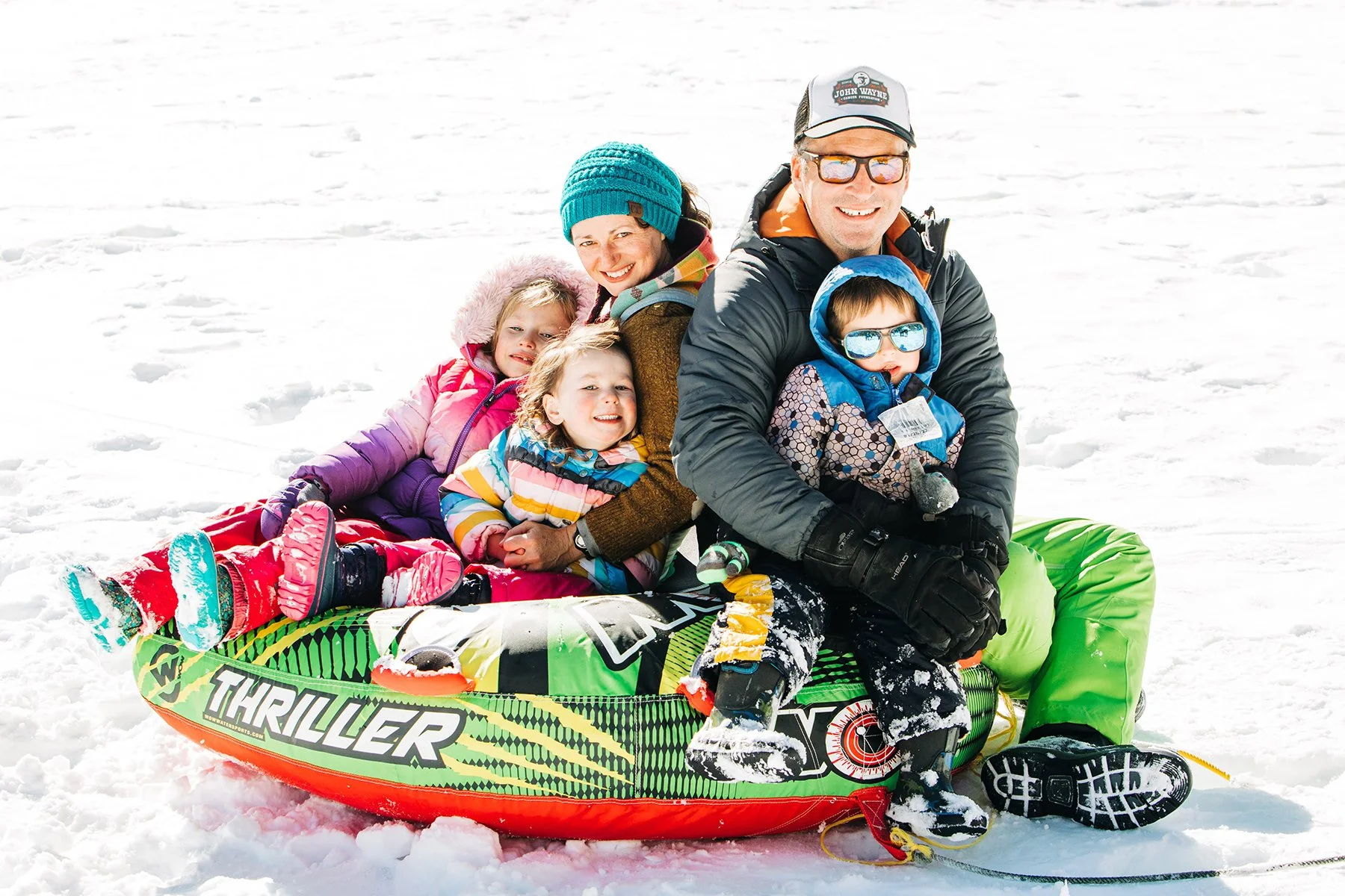 Family playing in snow on a sled, dressed in winter clothing with a sled labeled 'Thriller' in snowy landscape.