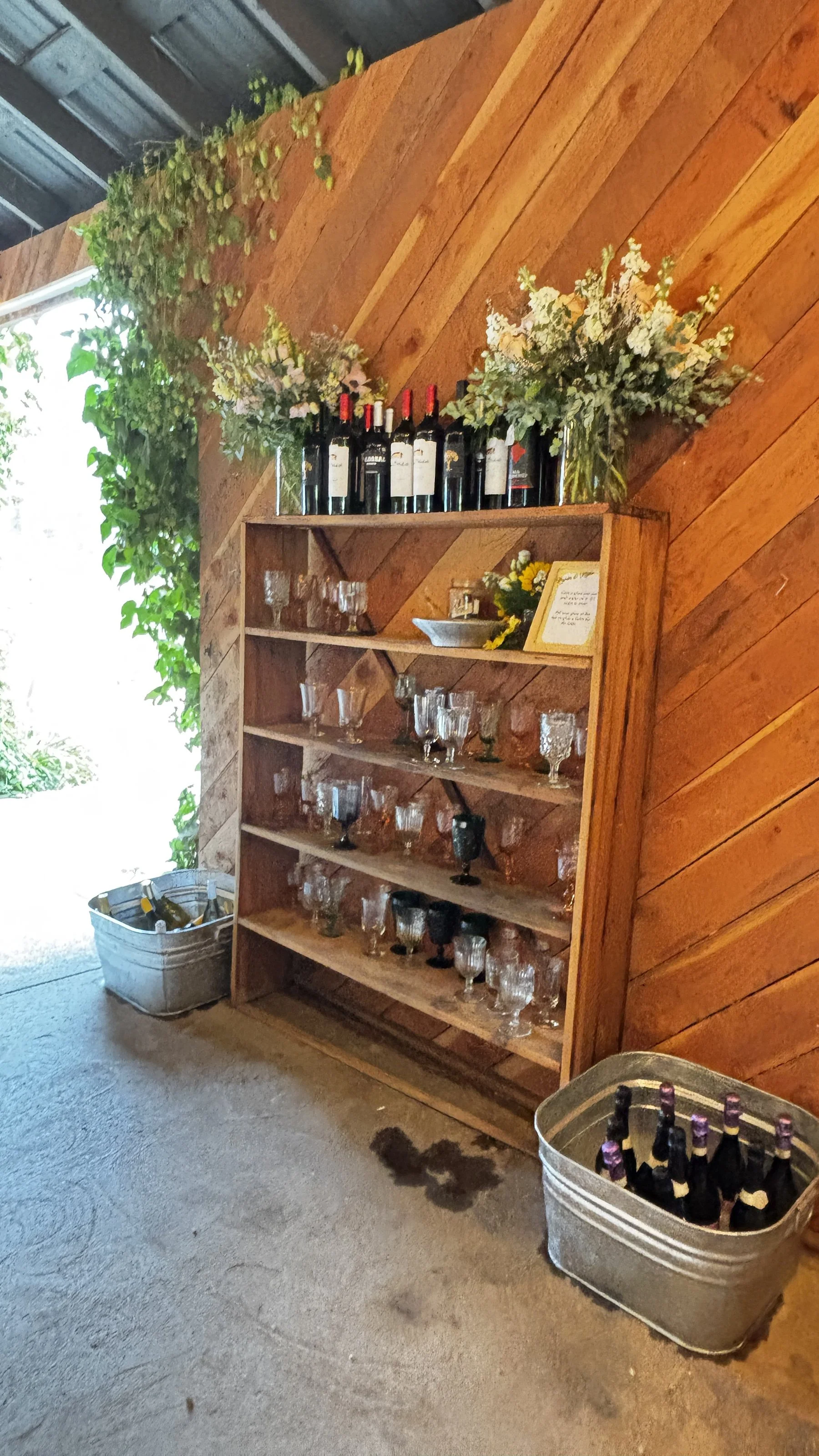 Wooden shelf holding wine bottles, glassware, and floral arrangements, with two metal containers of bottled drinks on a concrete floor.