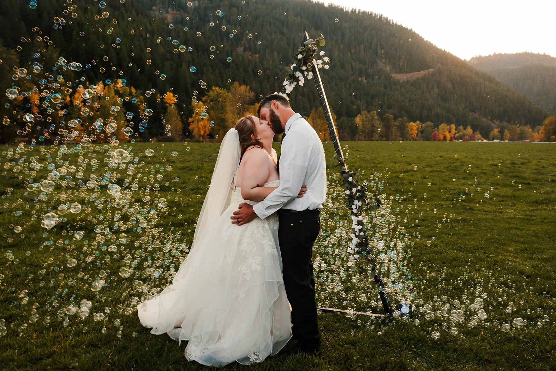 A bride and groom kissing outdoors in a grassy field at sunset, surrounded by bubbles and a mountain landscape in the background.