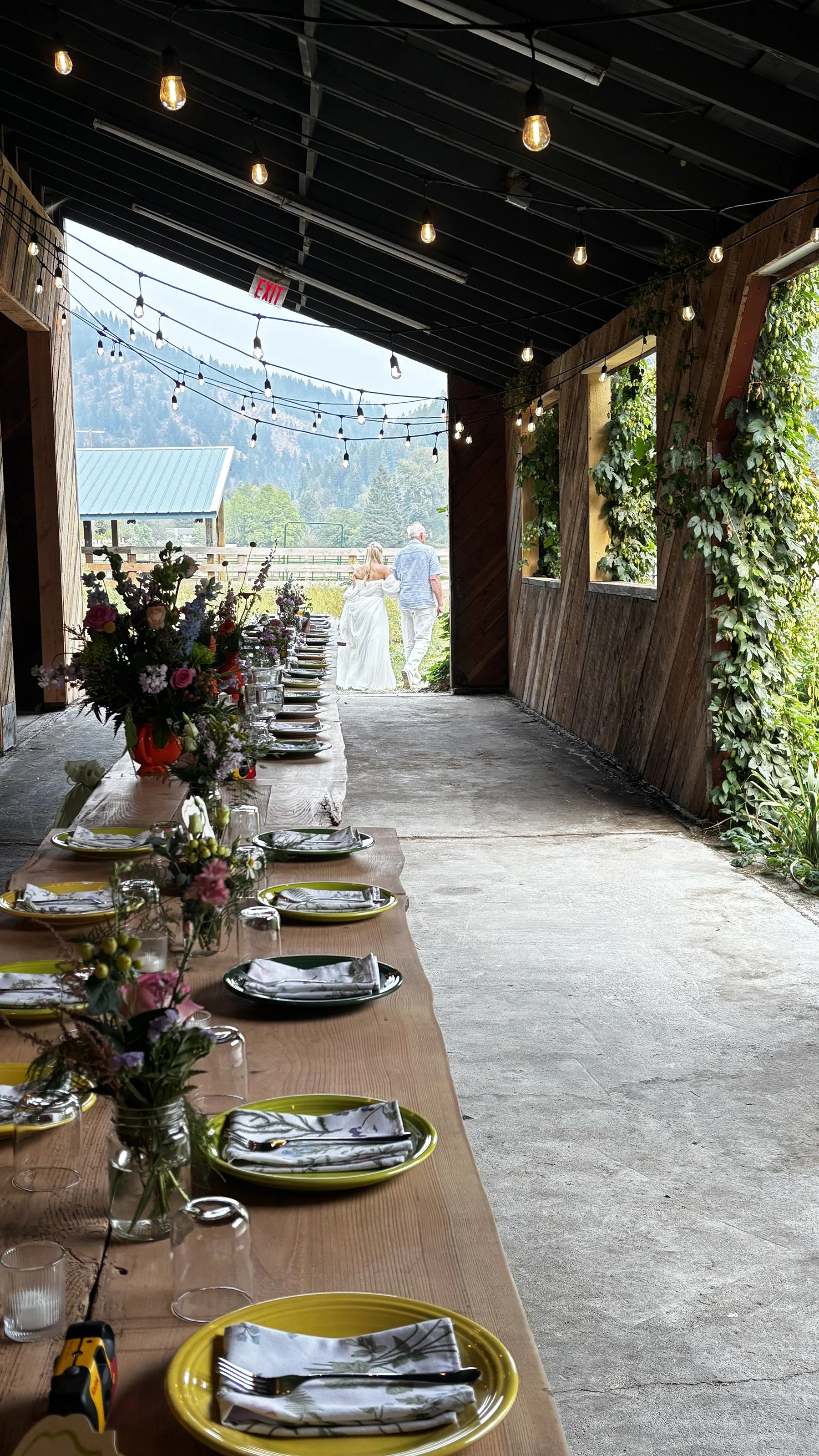 Long wooden table set with yellow plates, napkins, glasses, and flower arrangements, inside a rustic barn with string lights and open views of a field and mountains.