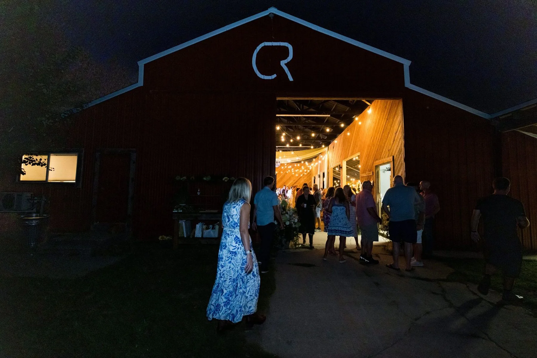 People entering a barn decorated with string lights for an event at night.