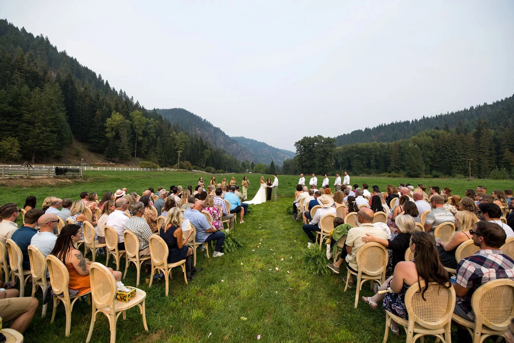 Outdoor wedding ceremony in a mountain valley with guests seated on chairs, officiant, bride, and groom at the altar surrounded by lush green trees and hills.