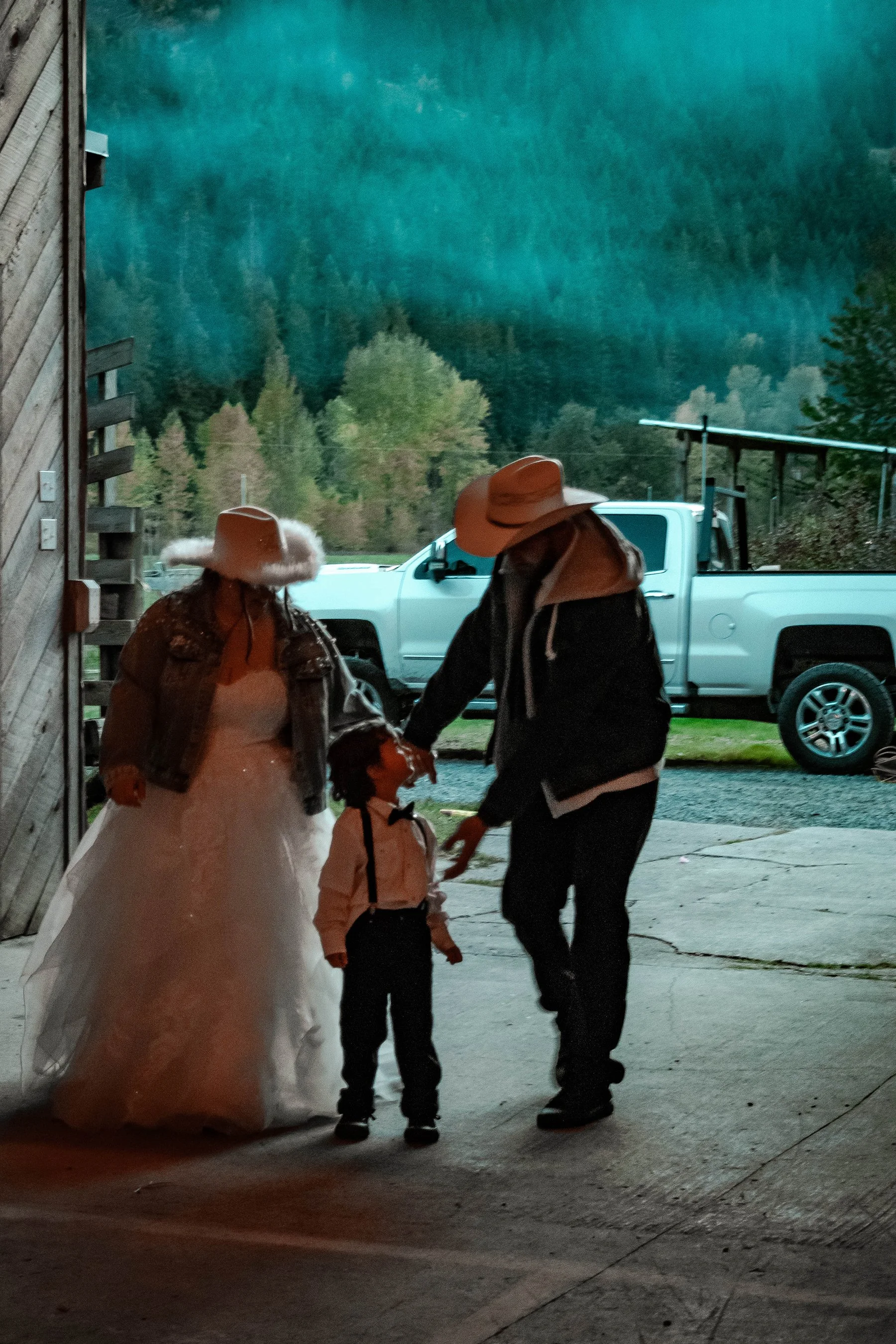 A woman in a wedding dress and a denim jacket, a man in a black jacket and cowboy hat, and a young boy in a white shirt and suspenders are holding hands and looking at each other outside near a white pickup truck, with a forested mountain background.