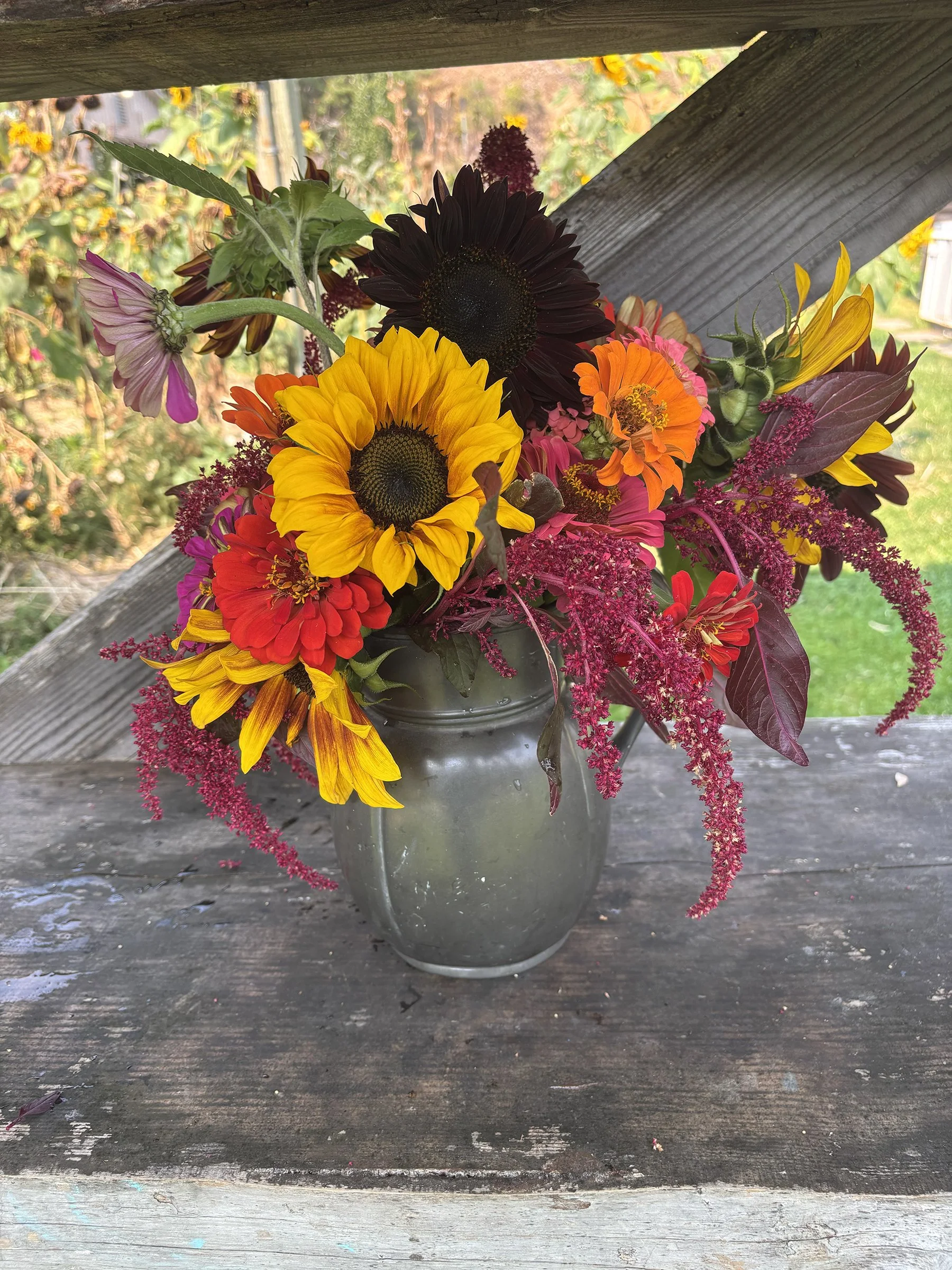 A colorful bouquet of sunflowers, zinnias, and other flowers in a gray vase on a weathered wooden surface, with a rustic outdoor background.