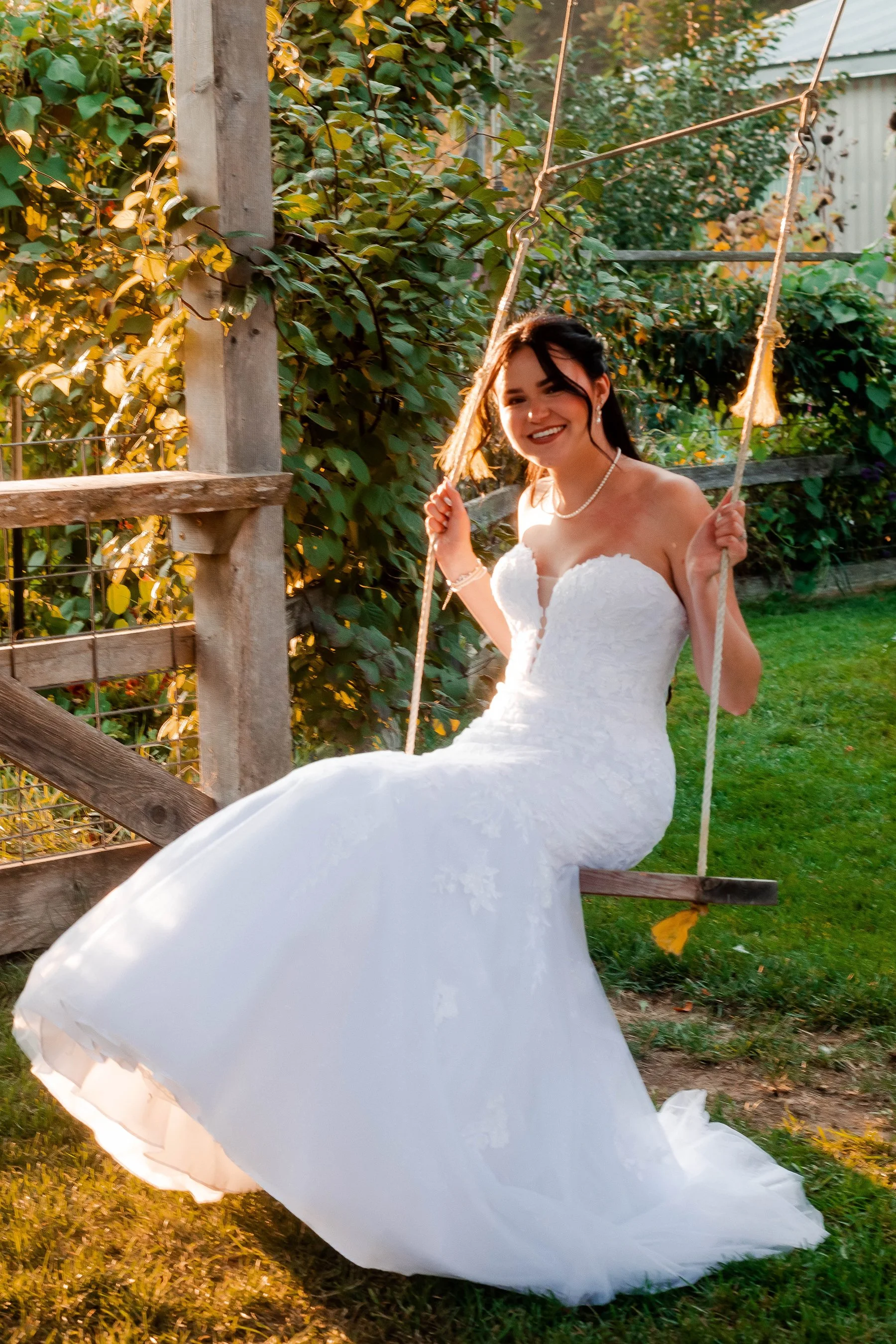 A bride in a white wedding gown sitting on a wooden swing outdoors during sunset.
