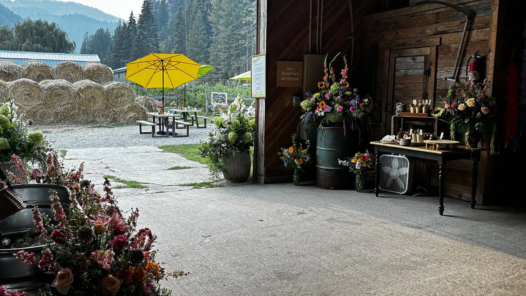 The interior of a rustic barn with floral arrangements and a table of craft supplies, with an outdoor seating area that has a yellow umbrella and hay bales in the background.