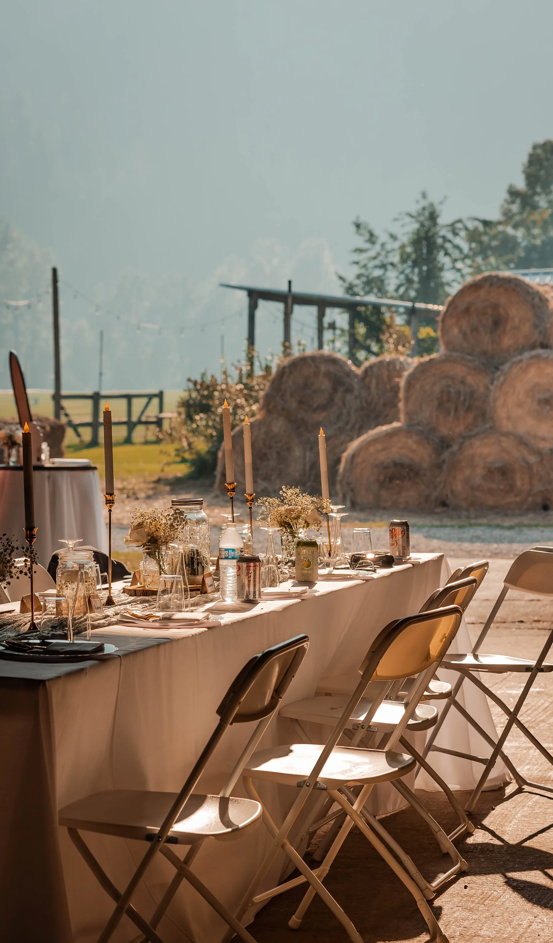 Long outdoor dining table set with white tablecloth, glassware, candles, and floral centerpieces, overlooking hay bales and a farm landscape during sunset.