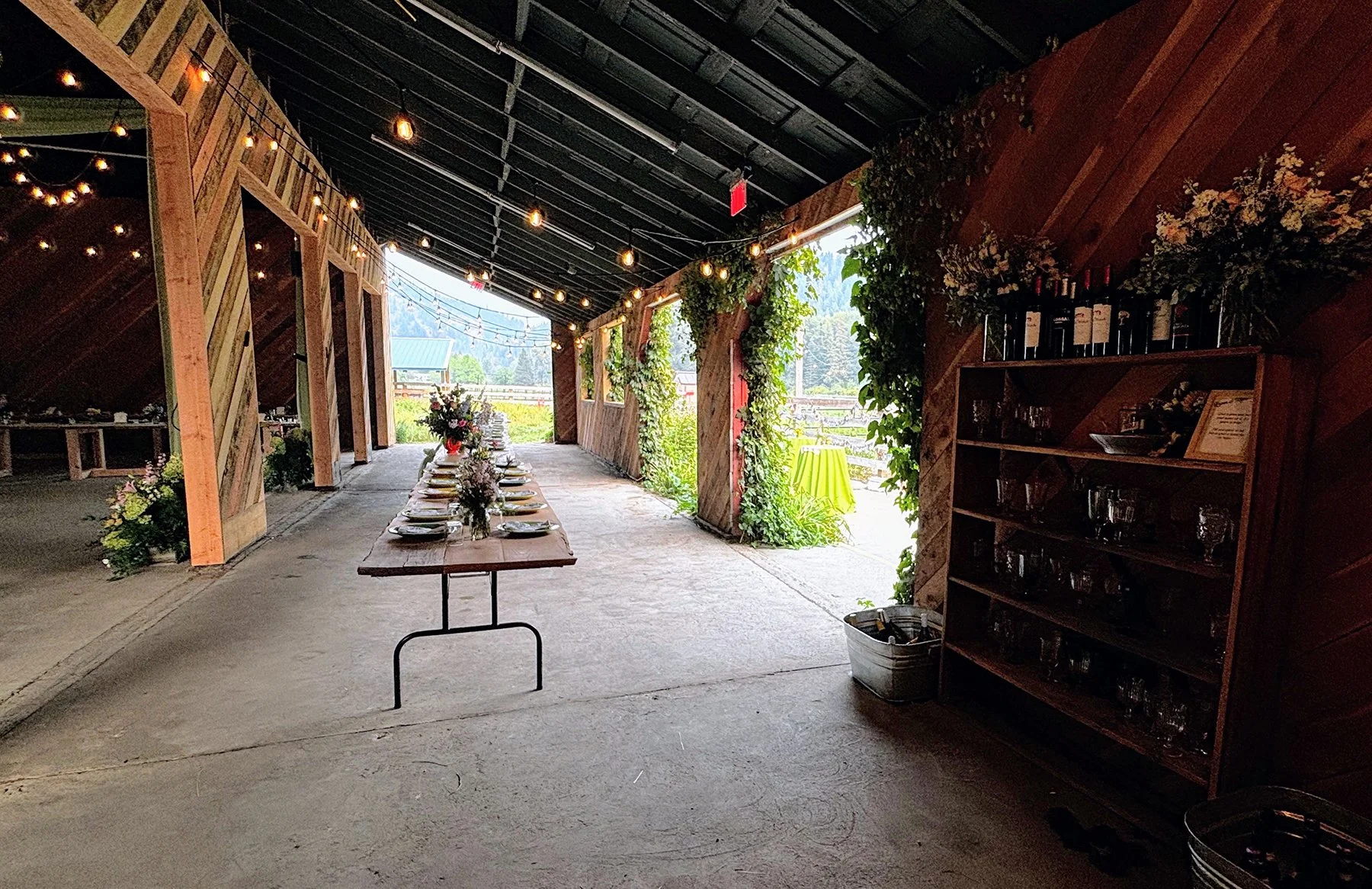 A rustic outdoor event space with a long table decorated with flowers and tableware, set under a covered wooden structure with string lights, surrounded by greenery and mountain views.