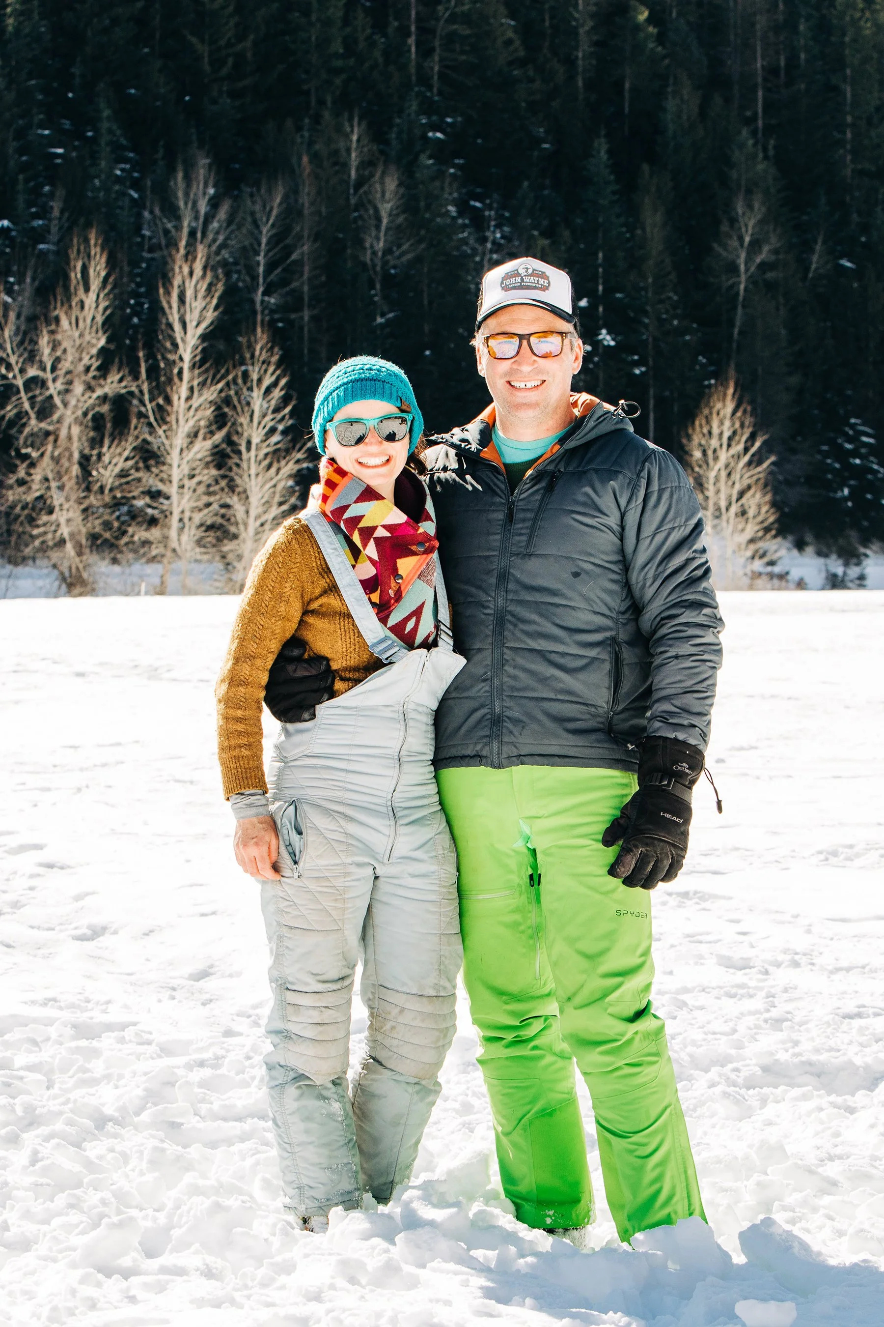 A smiling couple dressed in winter gear standing close together in a snowy landscape with a forest of snow-covered trees in the background.