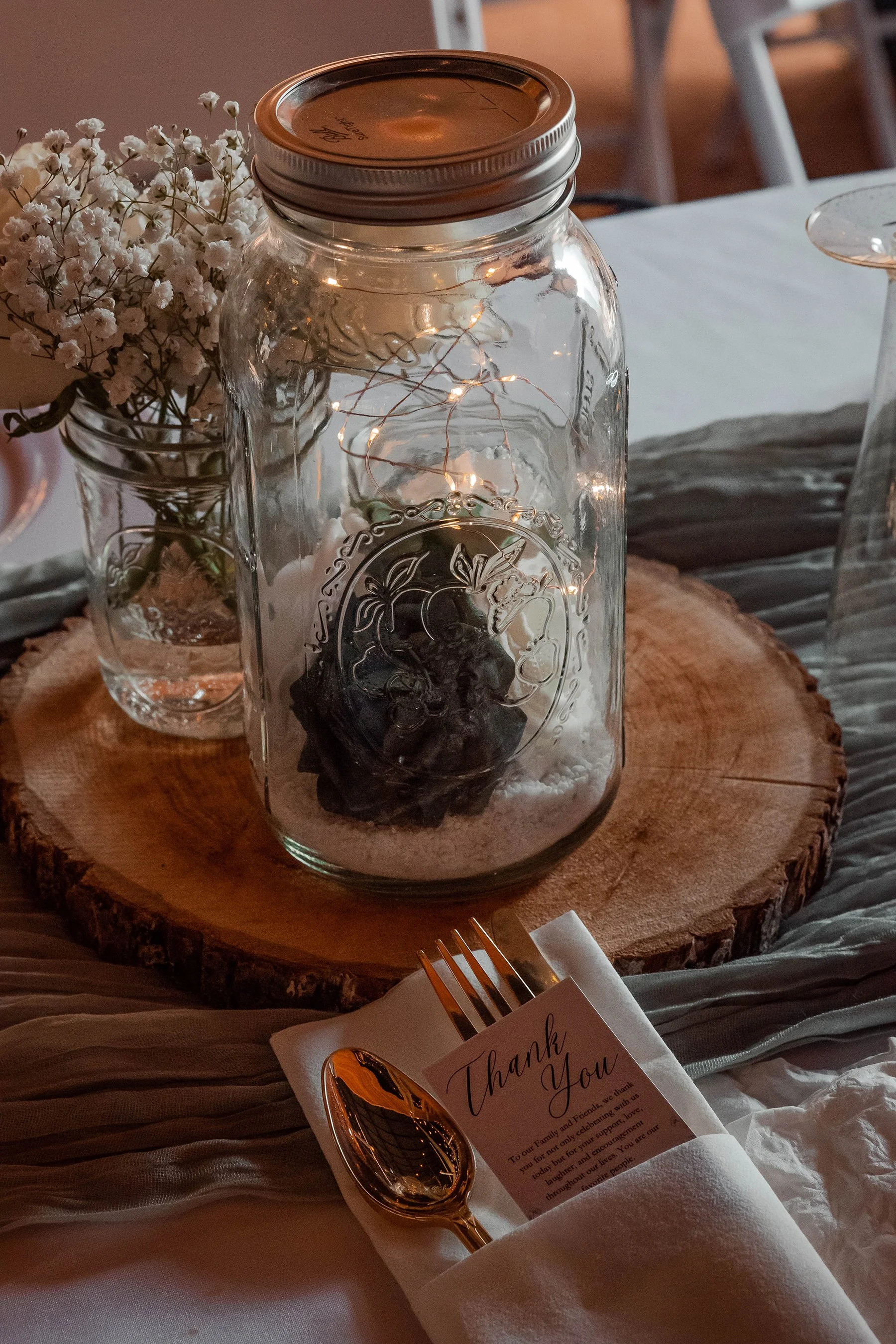 Decorative table arrangement featuring a glass jar with a sealed lid, illuminated fairy lights inside, and a black decorative item at the bottom. The jar is on a wooden slab with a white tablecloth underneath. To the left, there is a small vase with 