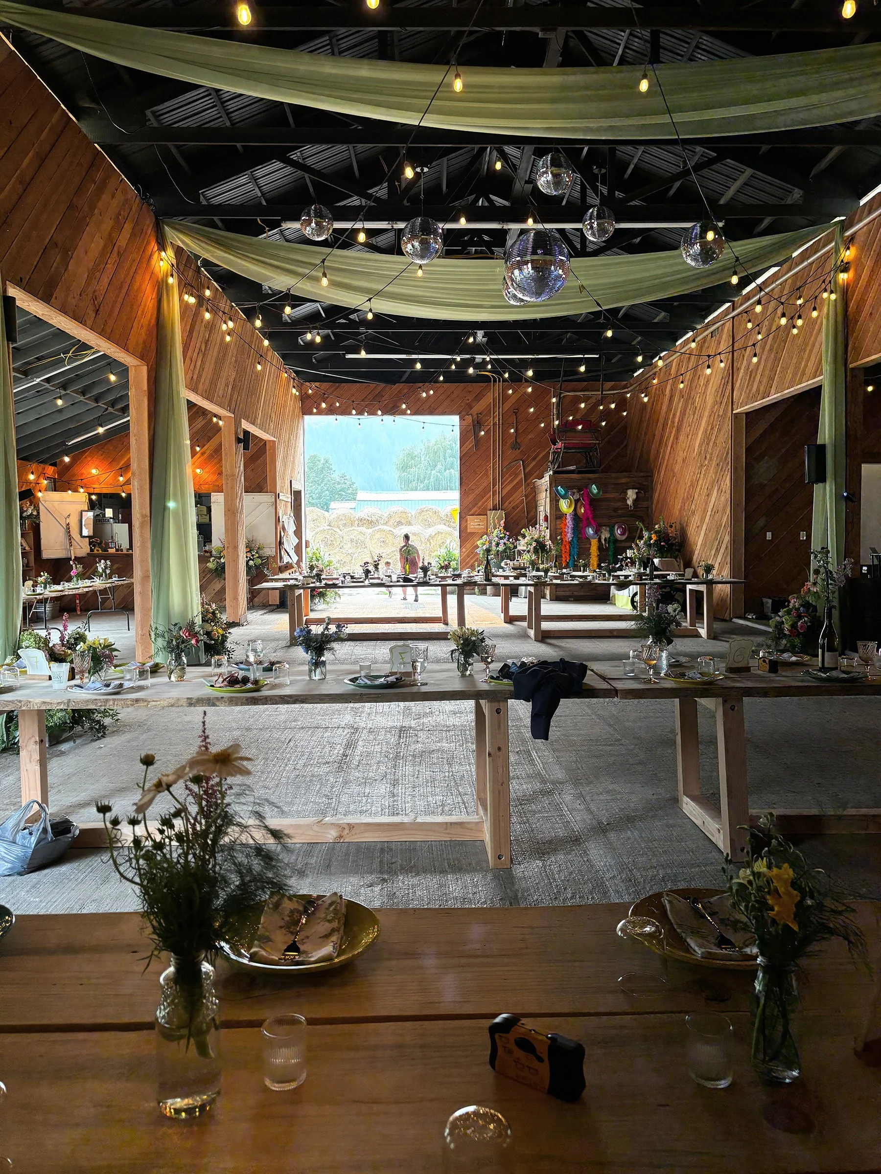 Decorated indoor event space with wooden walls, string lights, hanging disco balls, draped fabric, tables set with plates, glasses, and flowers, and a view of hay bales outside.