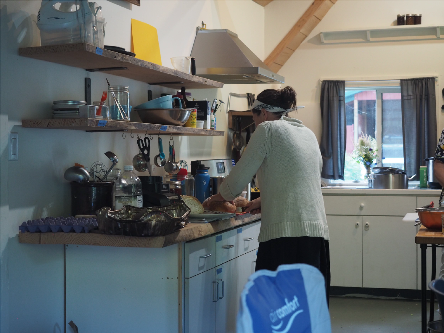 A woman in a sweater and headband preparing food on a kitchen countertop in a cozy, rustic kitchen with open wooden beams and a window with dark curtains.