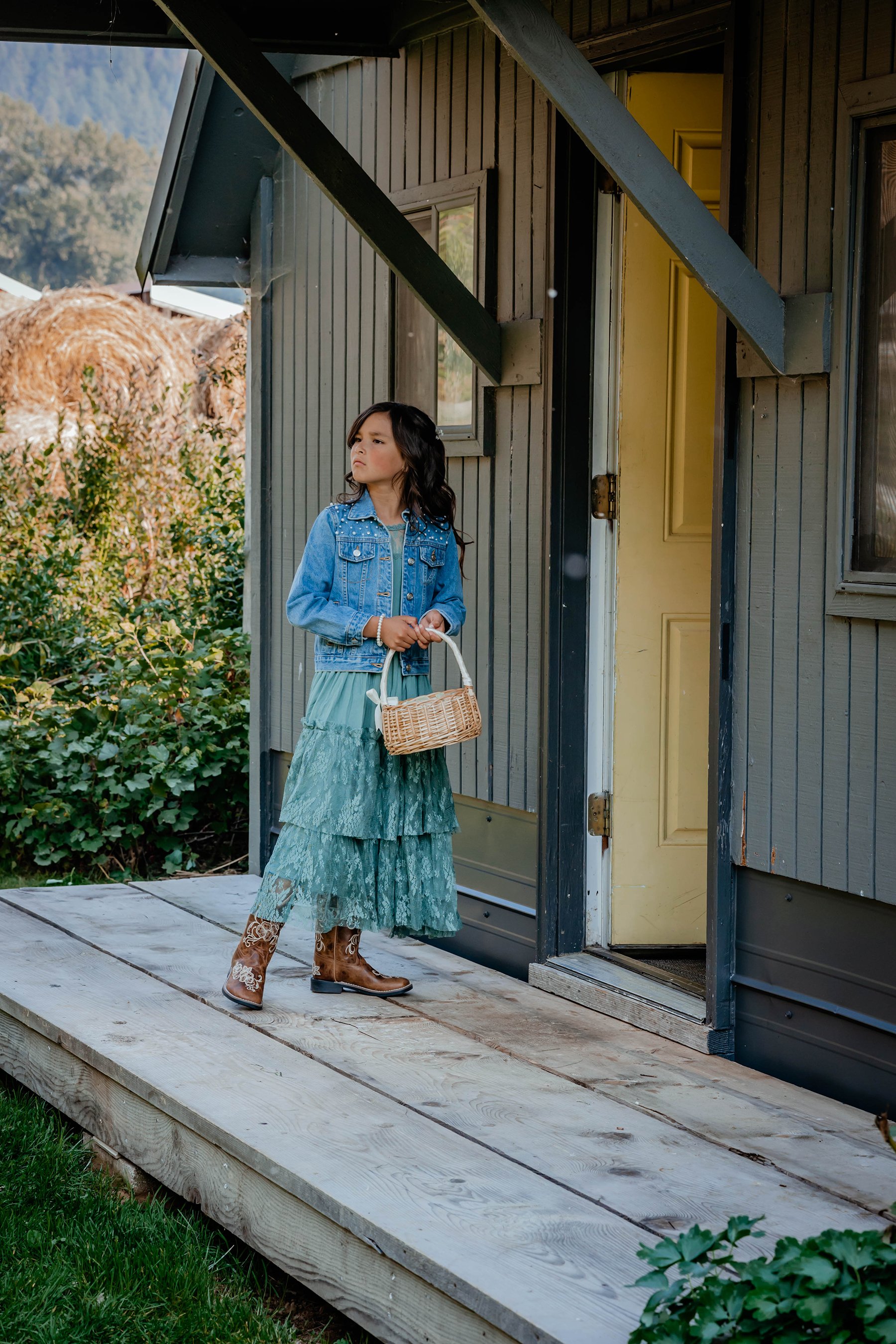 A young girl with long dark hair wearing a denim jacket and a teal layered dress, standing on a wooden porch holding a small wicker basket.