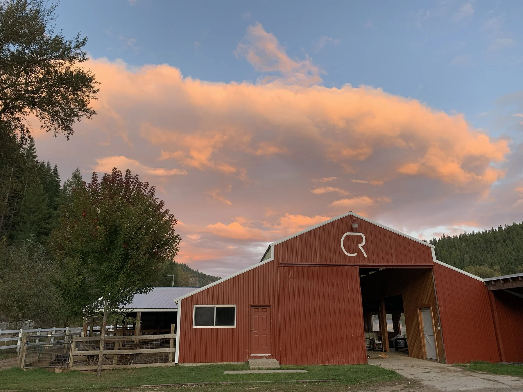 A red barn with an open door, surrounded by a fenced area and trees, under a colorful sunset sky with pink and orange clouds.