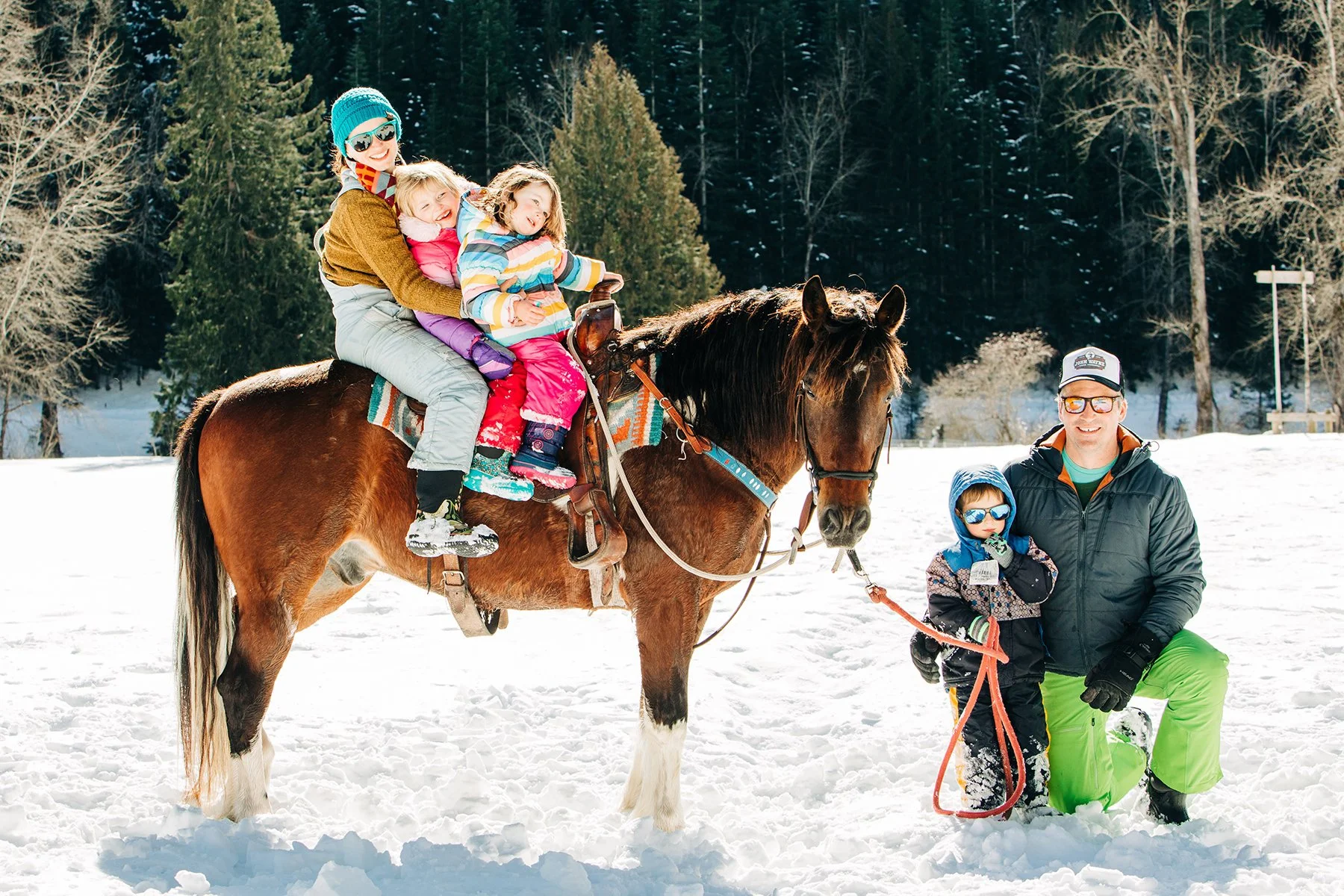 A family enjoying a winter outdoor activity in the snow. Several children are sitting on a horse, with one child being led by an adult male through the snow. The family is dressed warmly with colorful winter clothing, and the background features a fo
