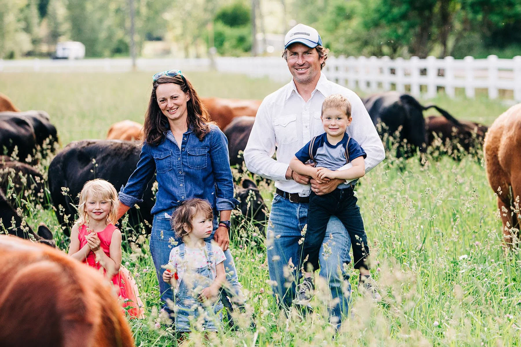 Family with three children on a farm, surrounded by cows in a grassy field, with a white fence and trees in the background.