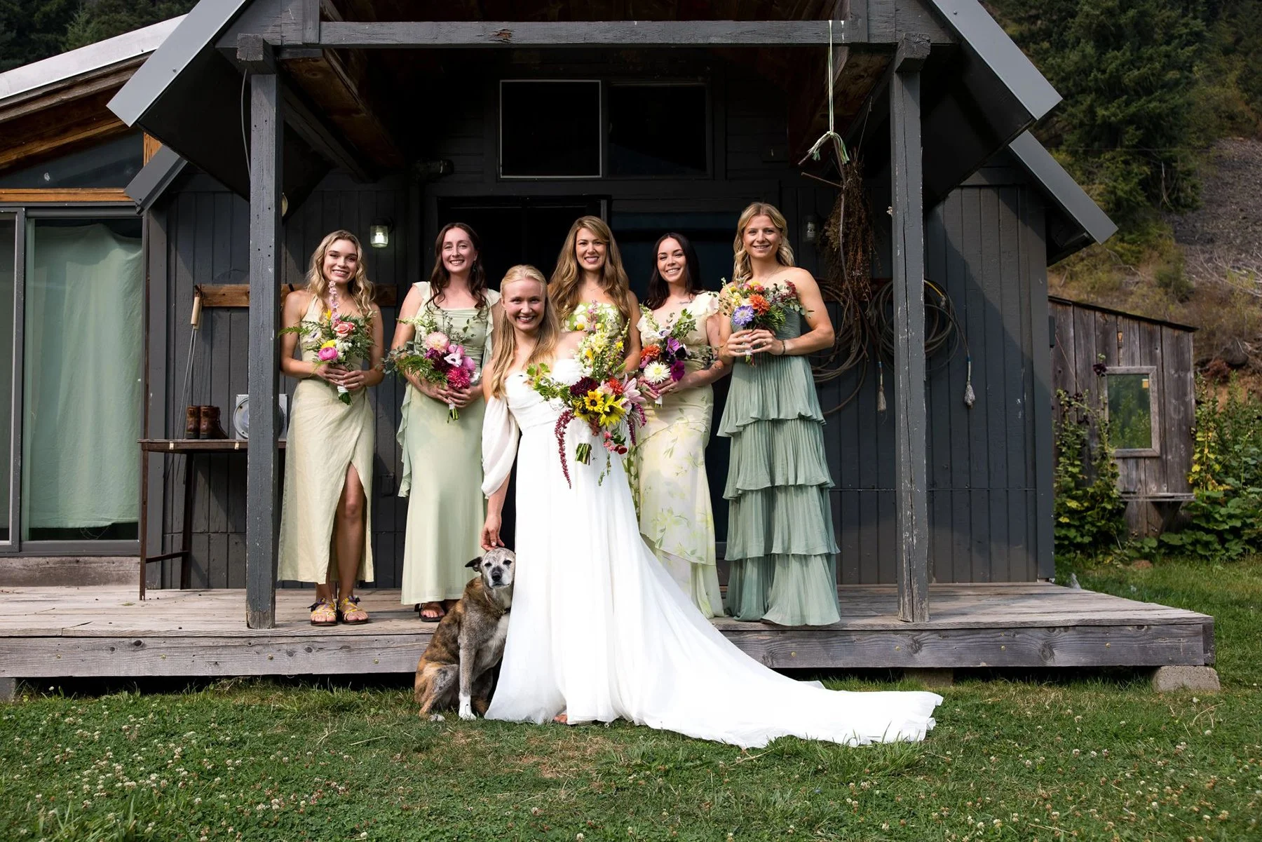 Group of six women, including a bride in a white wedding dress, standing on a wooden porch in front of a cabin, holding bouquets of flowers, with a dog sitting in front of the bride.
