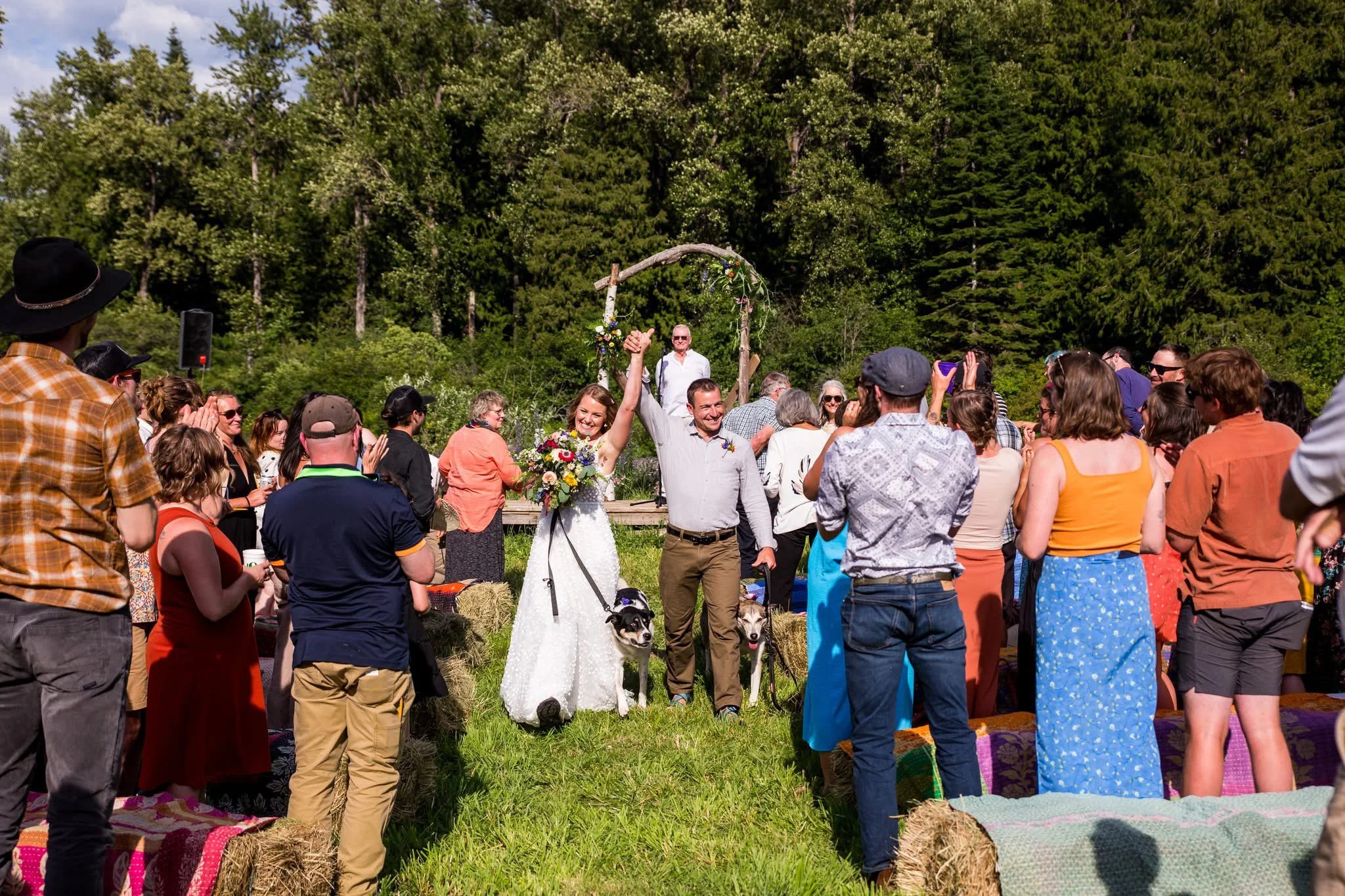 A newlywed couple celebrating at their outdoor wedding ceremony, holding hands with the bride holding a bouquet, surrounded by guests on a grassy field with hay bales, trees, and a decorative wedding arch in the background.