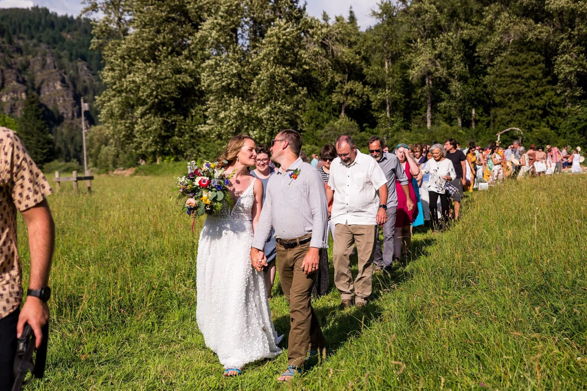 A wedding procession walking on a grassy field, with the bride in a white dress holding a bouquet of flowers, and people holding hands behind her, outdoors with trees and mountains in the background on a sunny day.