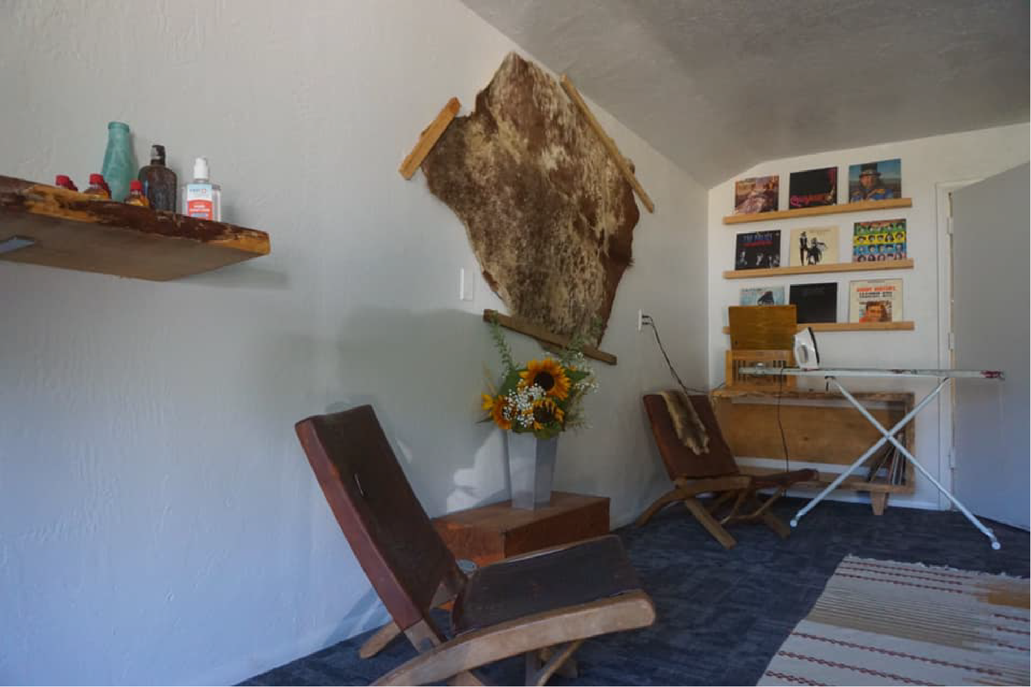 A cozy room with two wooden rocking chairs, a small wooden table, a decorative cowhide wall hanging, a built-in shelf with artwork and books, an ironing board, and a vase with sunflowers.