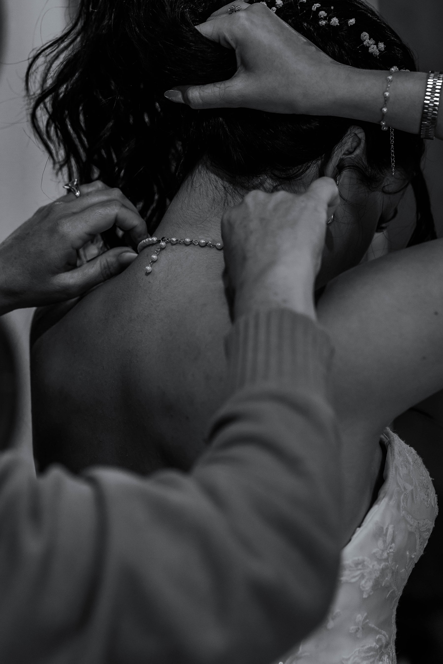 A woman with dark hair is being fitted with a pearl necklace by two other women. The image is in black and white and shows close-up hands adjusting jewelry around her neck.