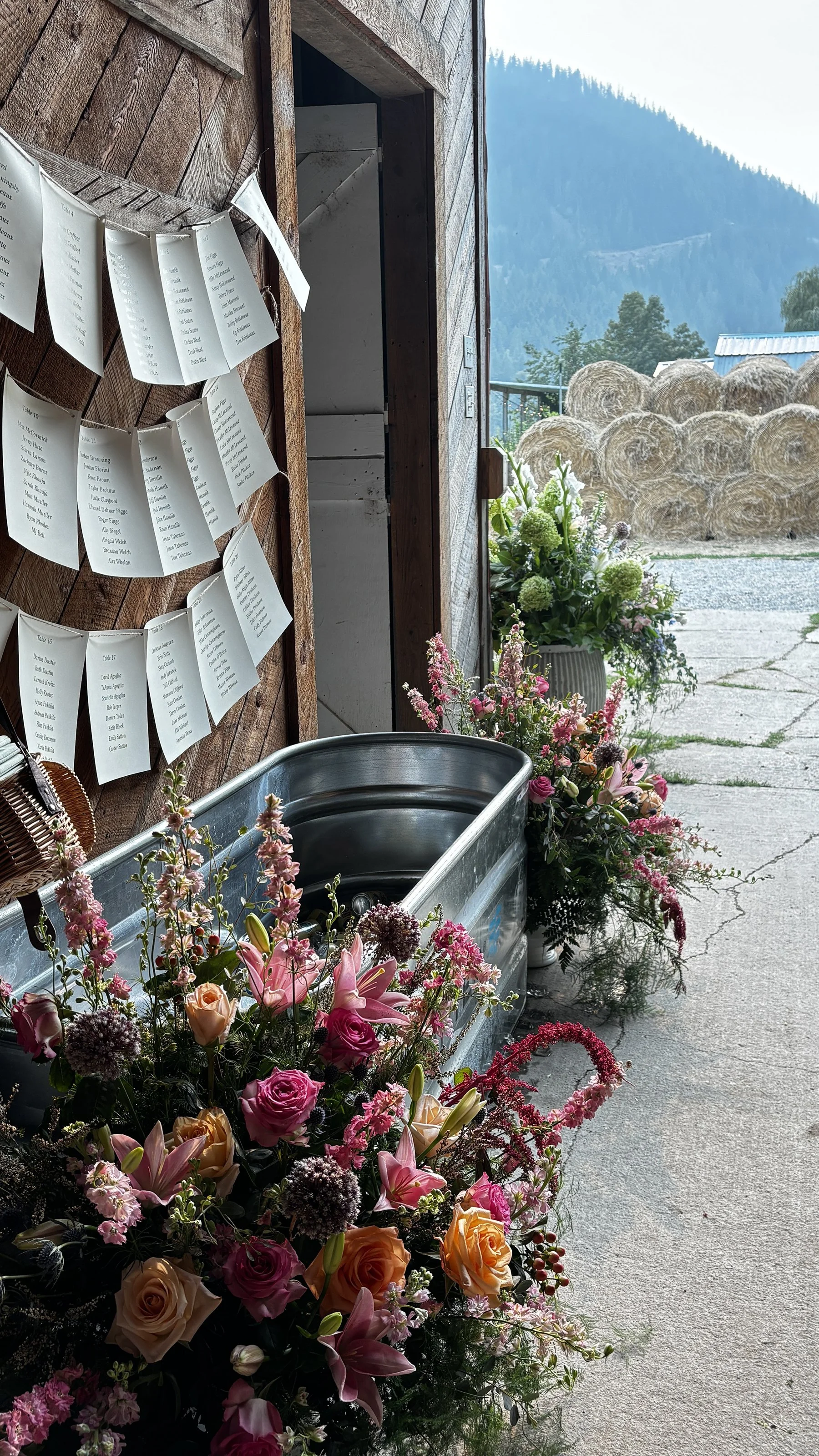 A rustic outdoor scene with a metal water trough filled with pink, white, and peach flowers, positioned on a concrete surface. To the right of the trough, there are more flower arrangements in vases. On the left, a wooden wall has sheets of paper han