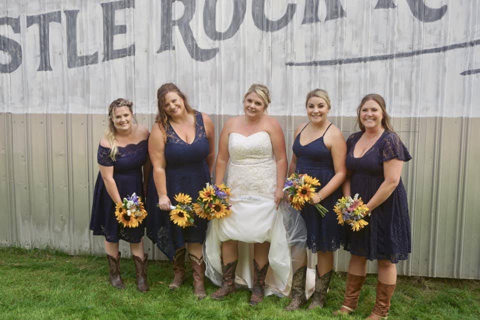 A bride and four bridesmaids in navy blue dresses with sunflowers, standing outdoors against a metal building wall. The bride is lifting her white dress and all are wearing cowboy boots.