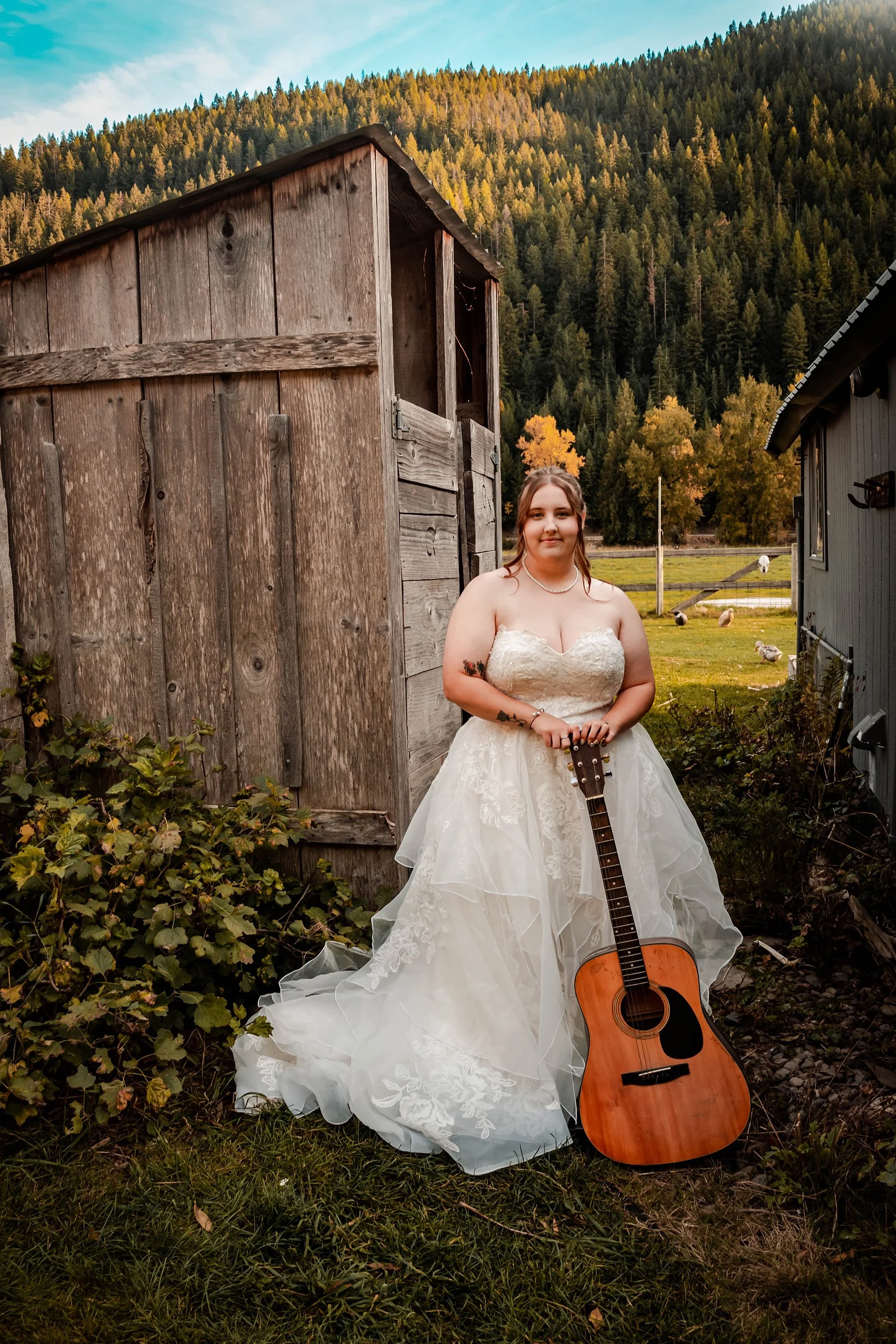 A bride in a white wedding gown holding a guitar standing outdoors near a rustic wooden building with trees and mountains in the background.