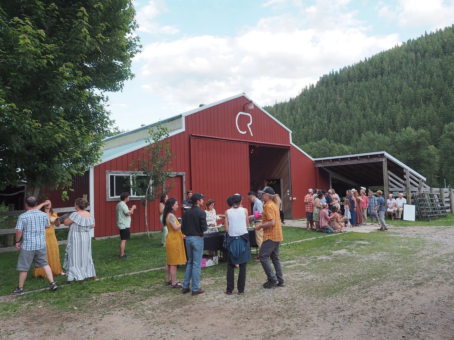 People gathering outside a red barn-style building with large group socializing and mingling on a grassy outdoor area, with green mountain hills in the background.