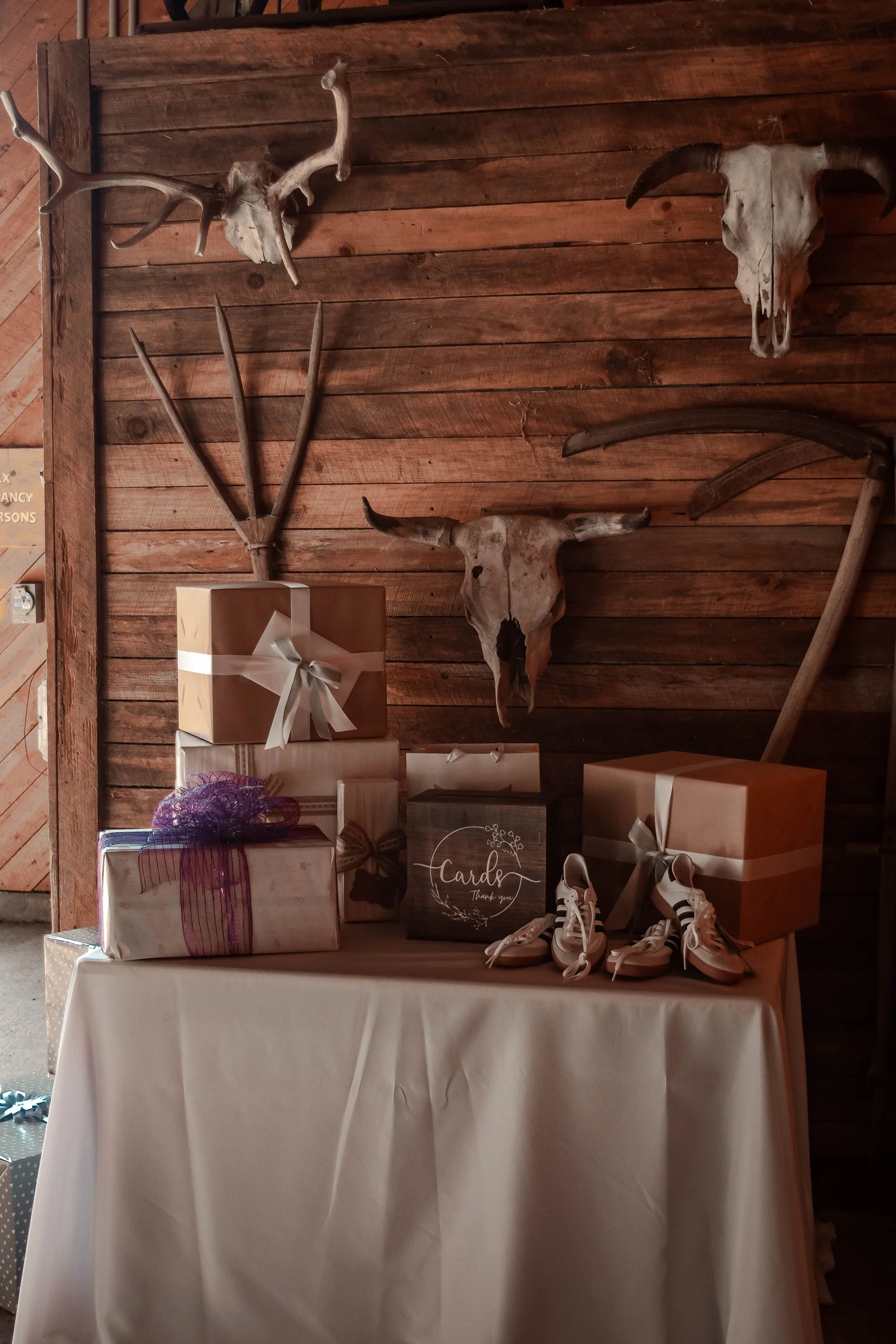 Gifts wrapped in brown and white paper, some with ribbons, placed on a white table with a wooden wall background decorated with mounted animal skulls and antlers, and a pair of white sneakers.