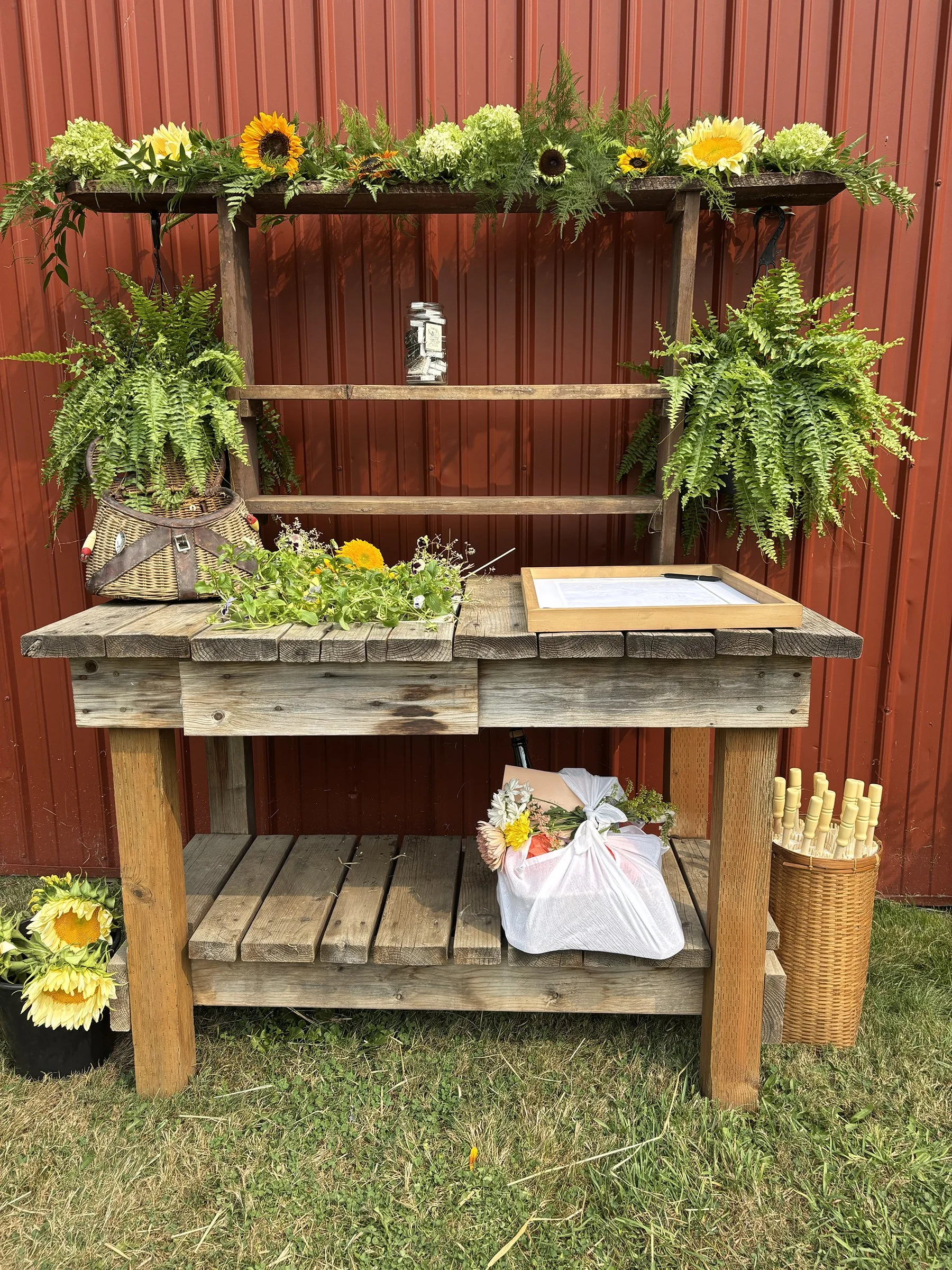 A rustic wooden outdoor display stand with greenery, sunflowers, and flowers, set against a red metal wall.