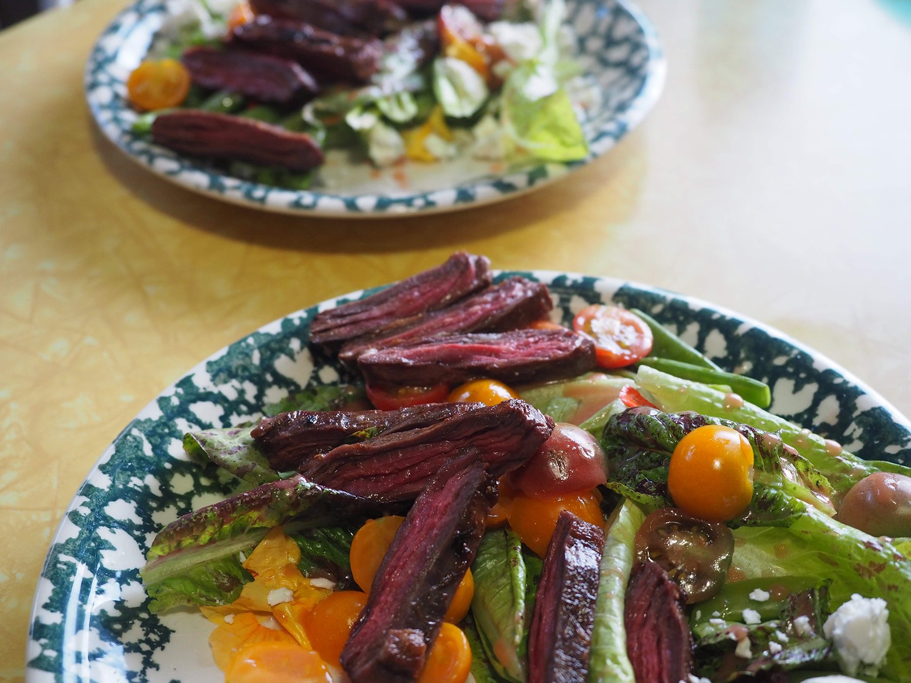 Close-up of a fresh salad on a patterned plate with mixed greens, cherry tomatoes, toasted bread slices, and feta cheese. Another plate with similar salad is visible in the background.