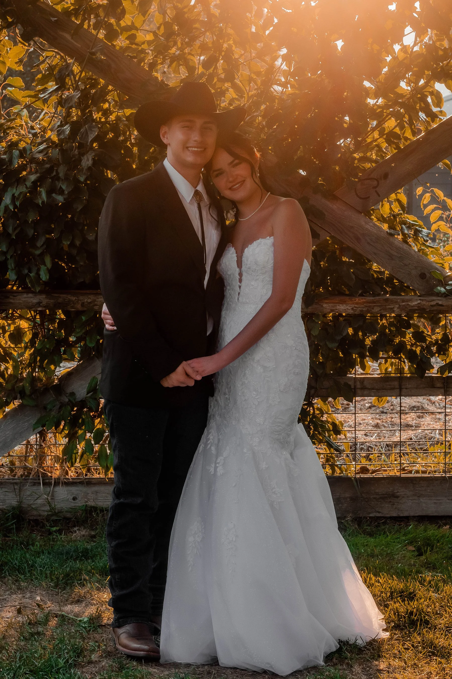 A bride and groom holding hands and smiling outside at sunset, with trees and a wooden fence in the background.