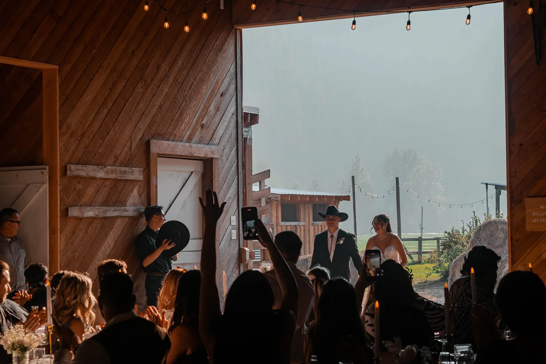 A bride and groom walk into a rustic barn wedding reception, with guests seated and taking photos, some raising their hands, and outdoor scenery visible through the barn's large open doorway.