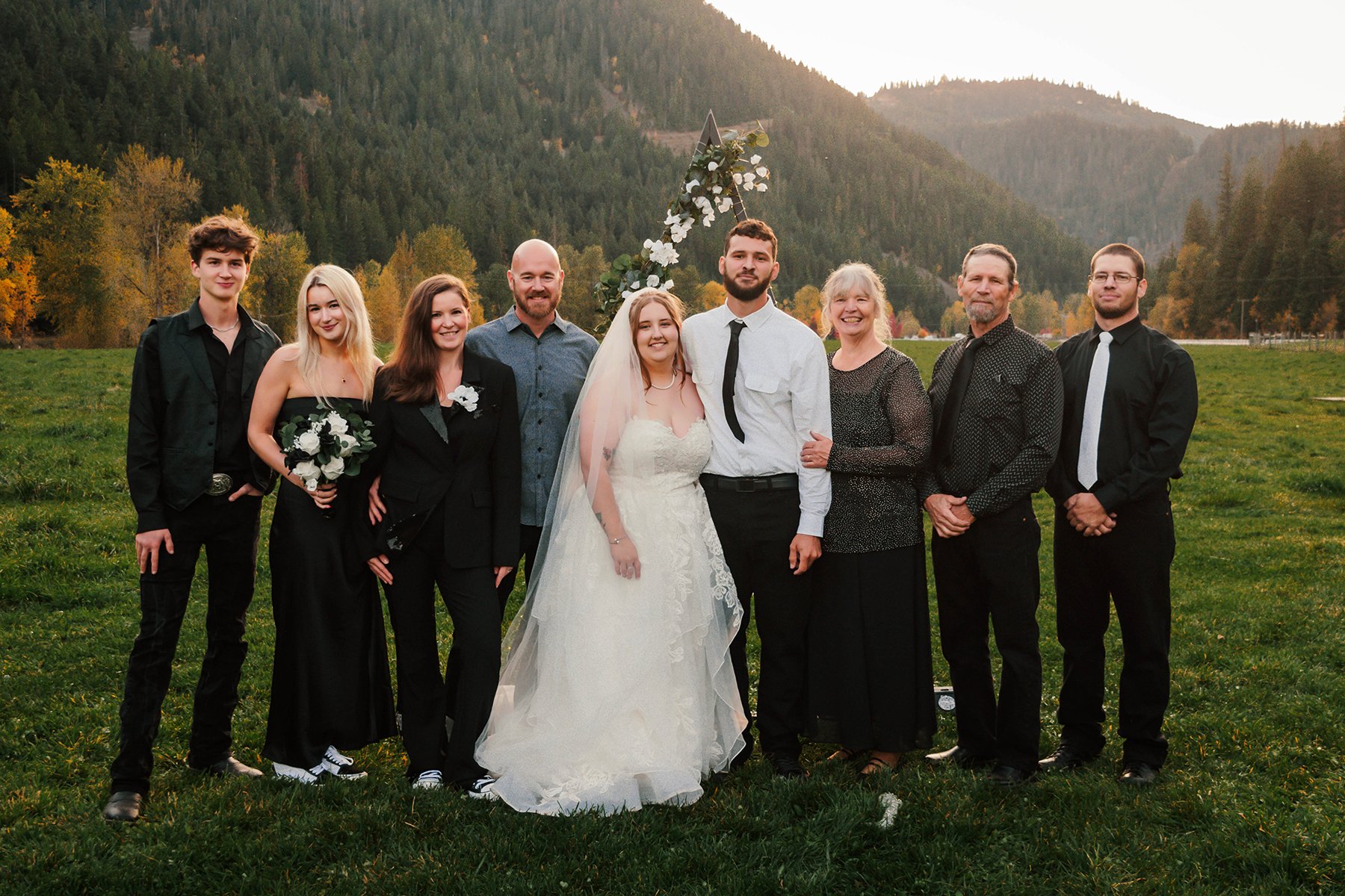 A group of nine people, including a bride and groom, standing outdoors in a grassy field with mountains and trees in the background during sunset.