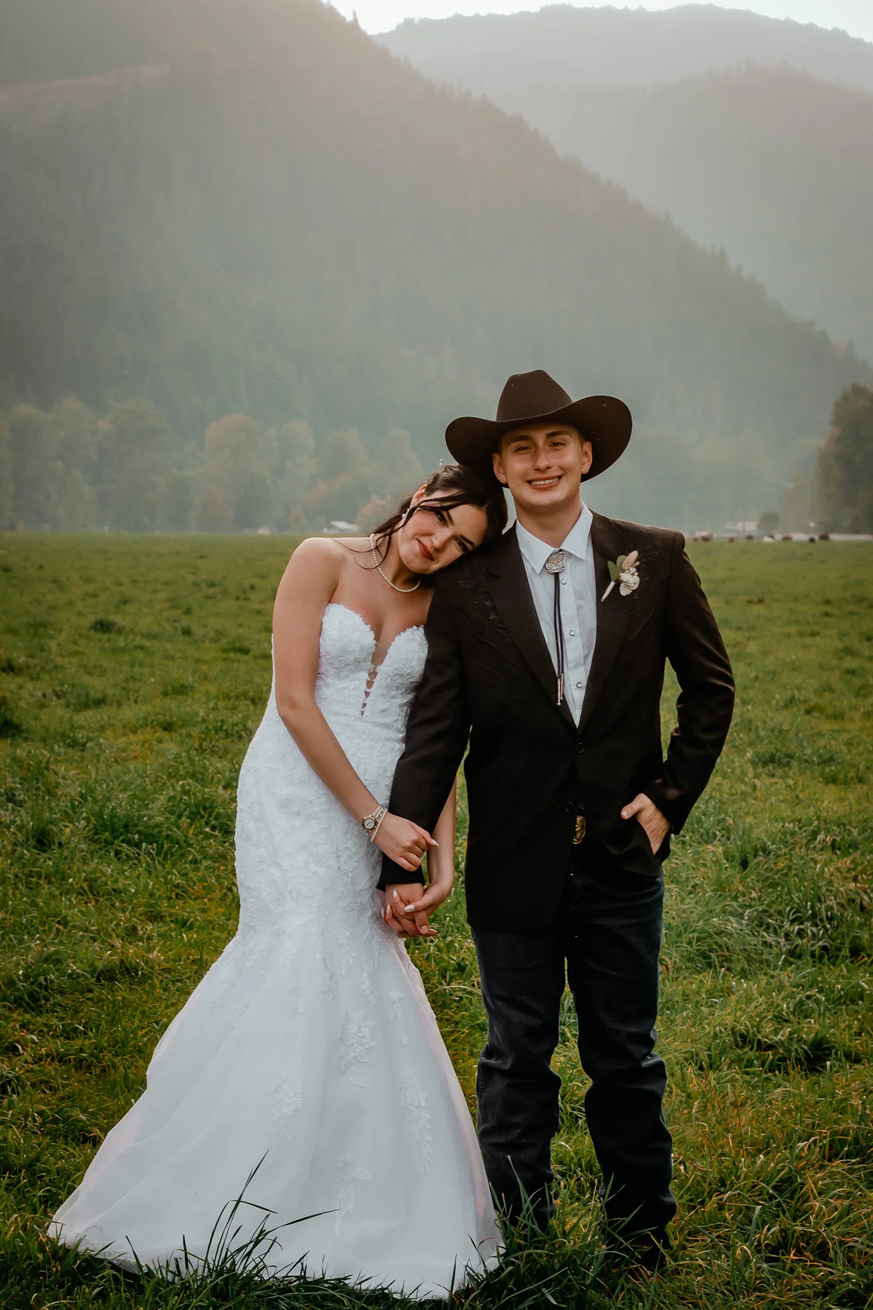 A bride and groom standing in a grassy field with mountains in the background, smiling and holding hands. The bride is in a strapless white wedding dress, and the groom is wearing a black cowboy hat and a black suit with a bolo tie.