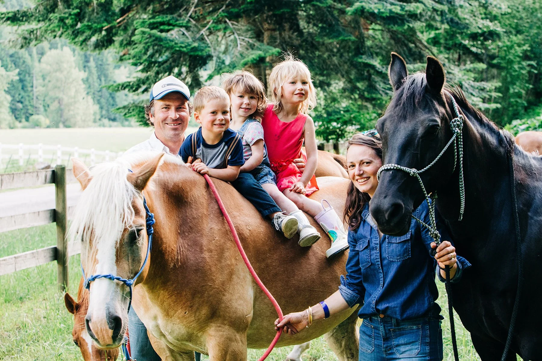 A family enjoying a pony ride outdoors, with three children sitting on a brown pony and two adults holding the pony's reins, surrounded by green trees.