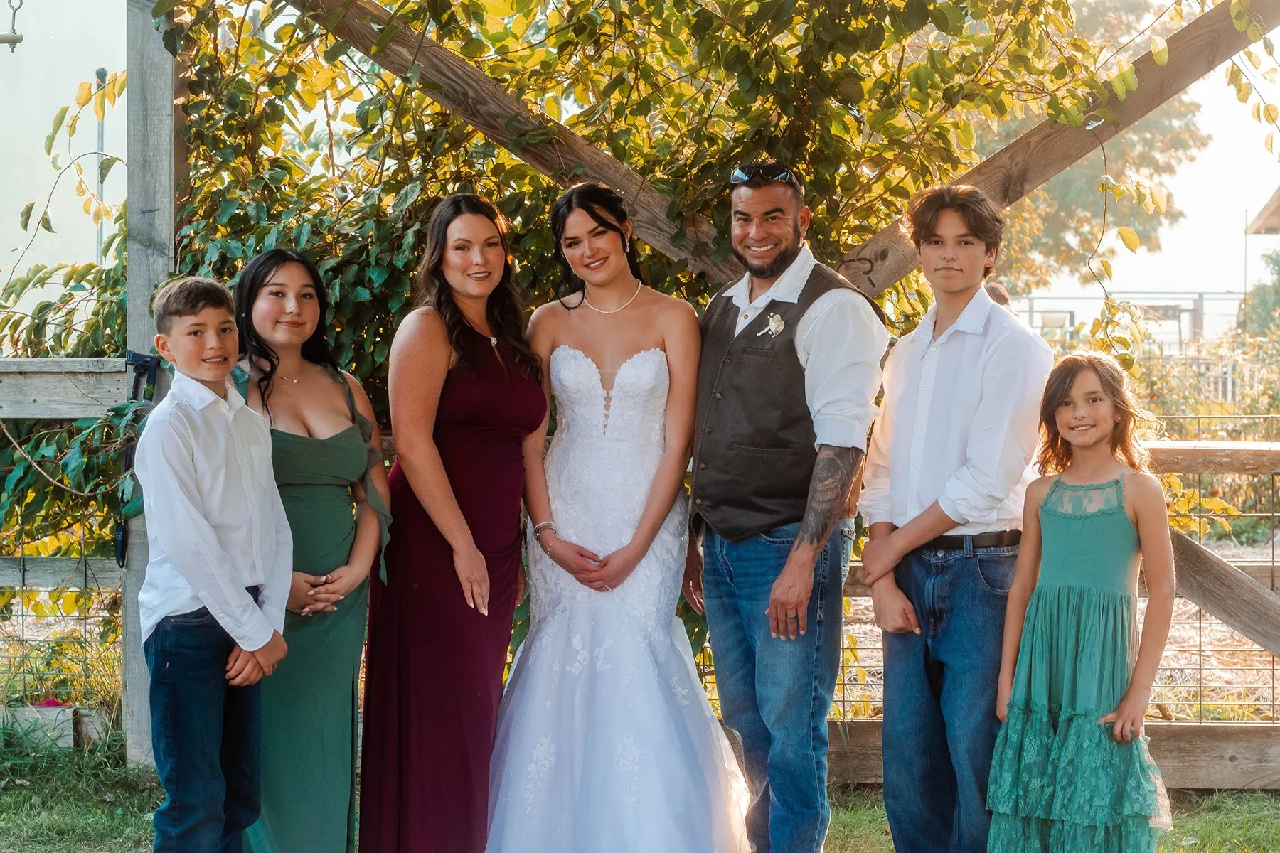 Group of seven people, including a bride in a white wedding dress, a groom in a vest, two women, and four children, standing outdoors in front of lush green trees and a wooden fence.