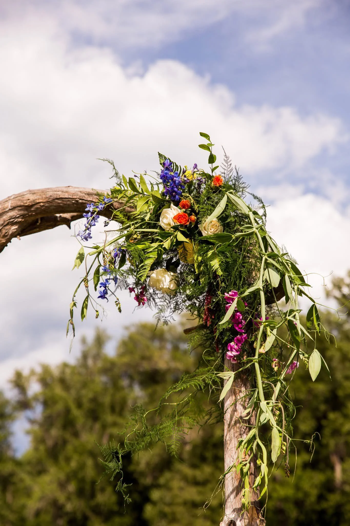 A decorative floral arrangement attached to a wooden arch, featuring various colorful flowers and green foliage against a cloudy sky.