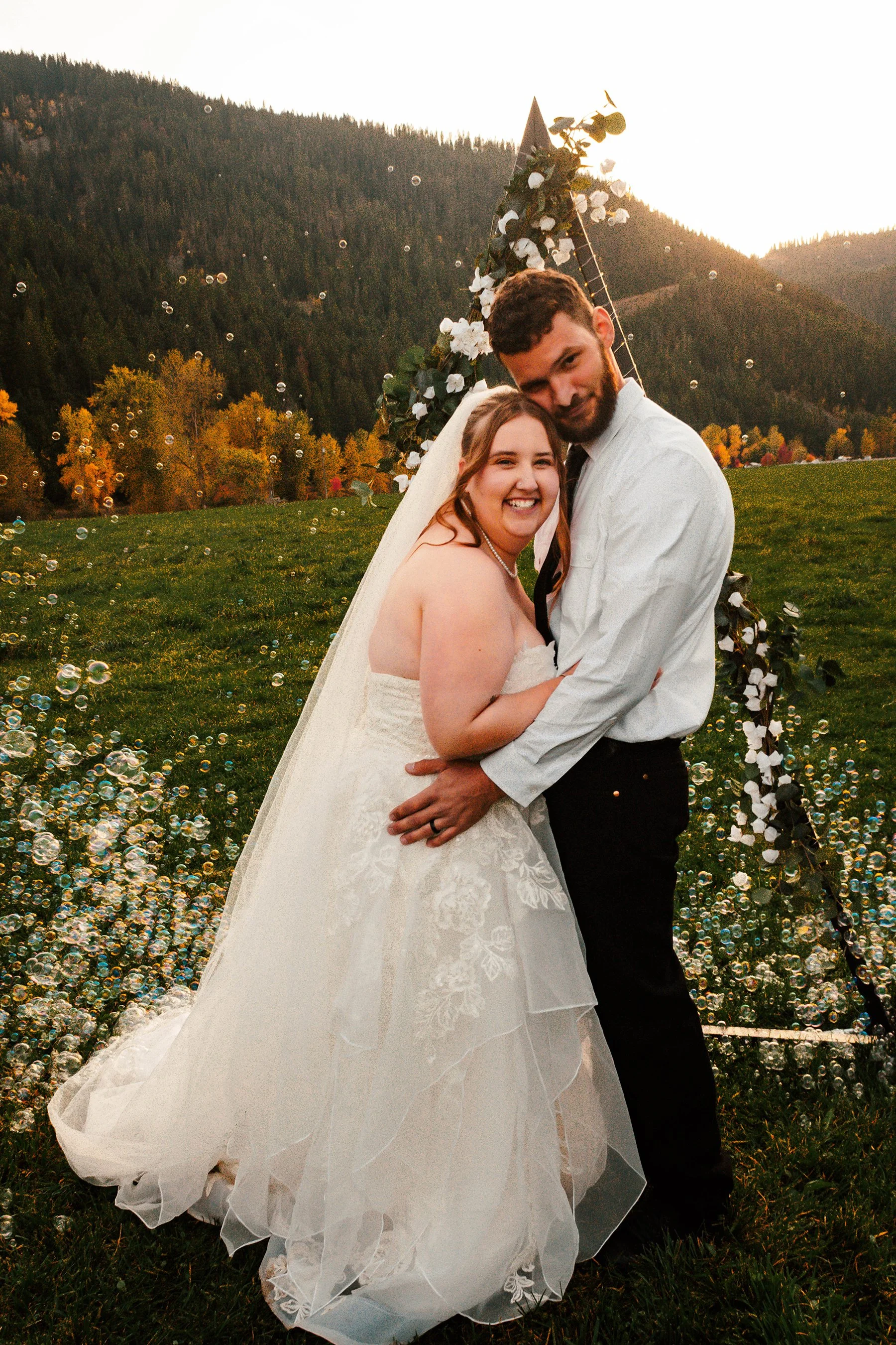 A newlywed couple smiling and embracing outdoors during sunset with a scenic mountain background, decorated wedding arch, and bubbles floating in the air.